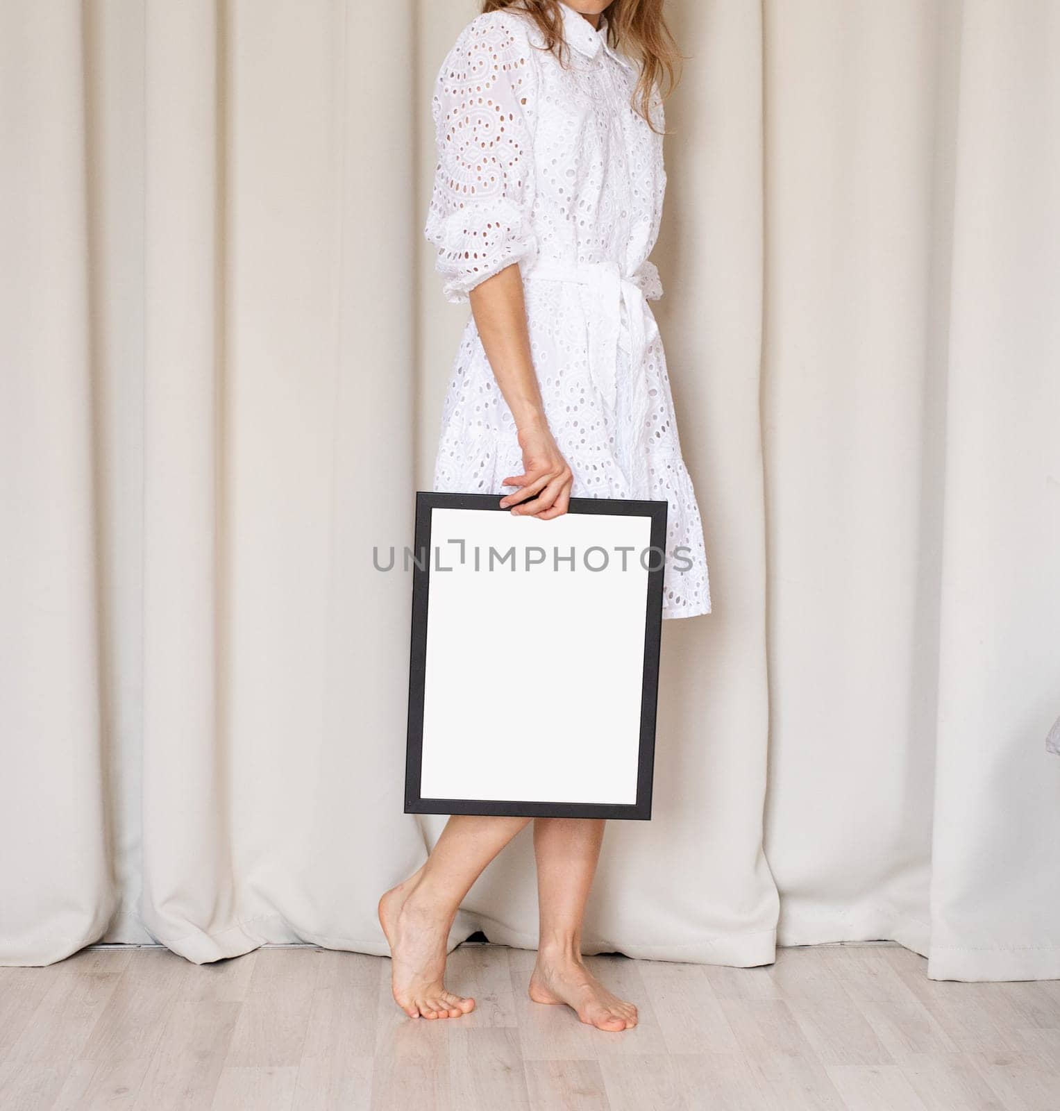 unrecognizable barefoot woman in beautiful white dress holding blank frame, beige curtain background, mockup design