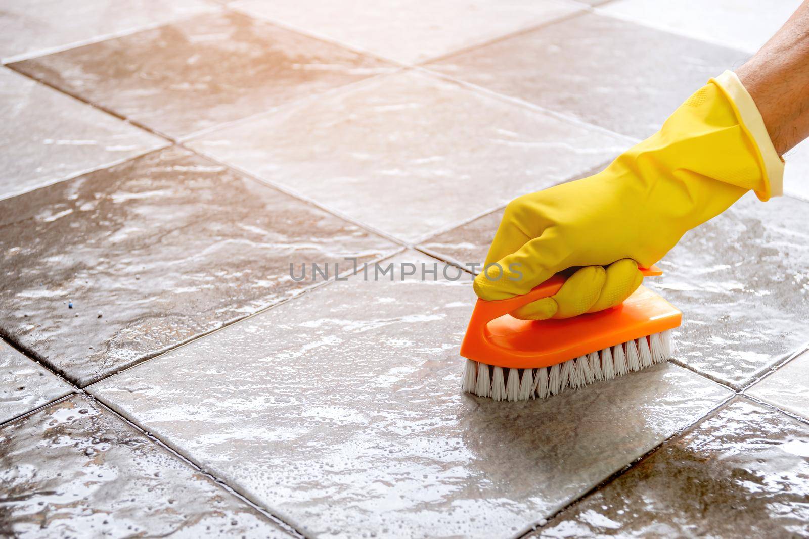 Hands wearing yellow rubber gloves are using a plastic floor scrubber to scrub the tile floor with a floor cleaner. by wattanaphob