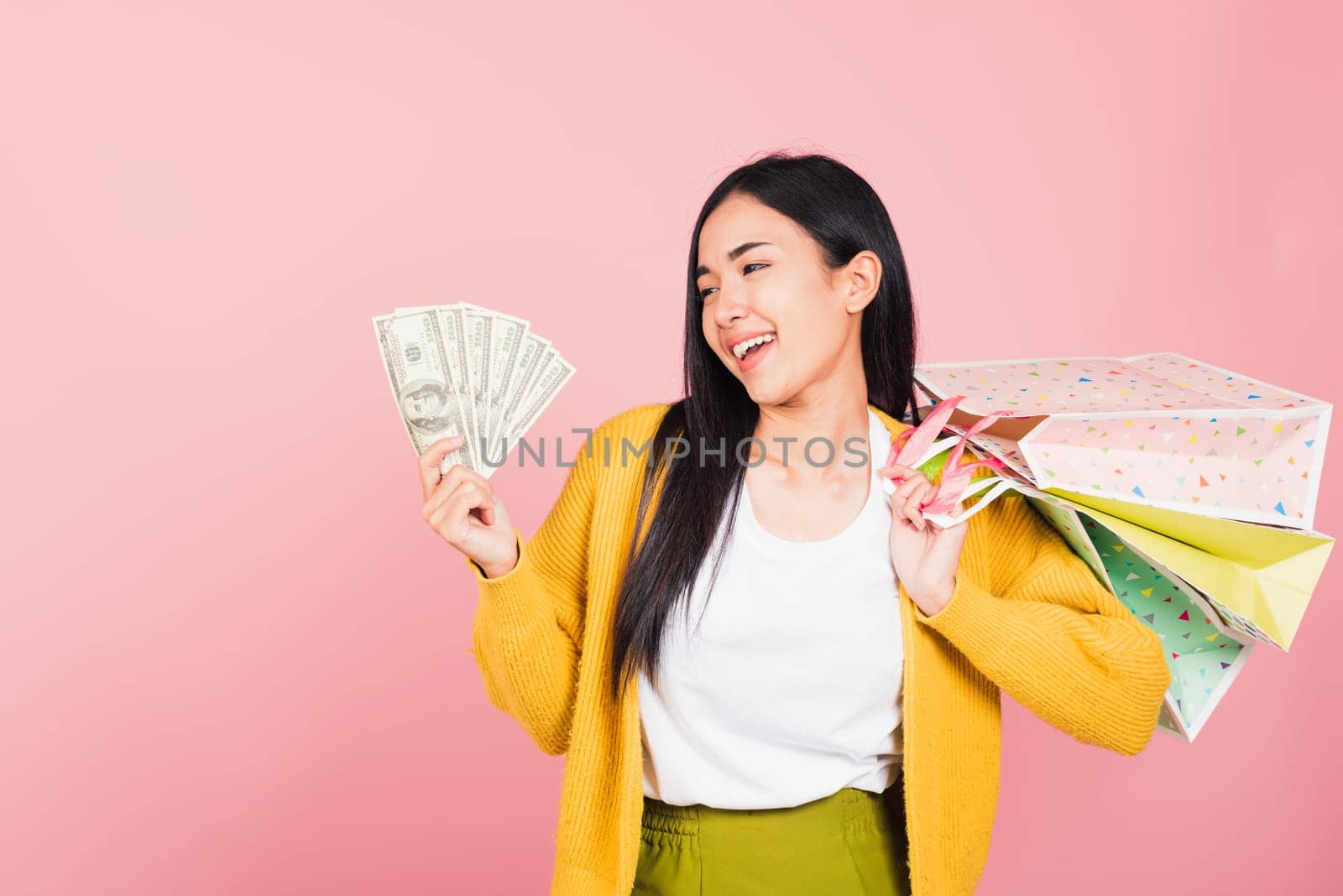 Portrait Asian happy beautiful young woman shopper smile excited holding online shopping bags colorful multicolor and dollar money banknotes on hand in summer, studio shot isolated on pink background