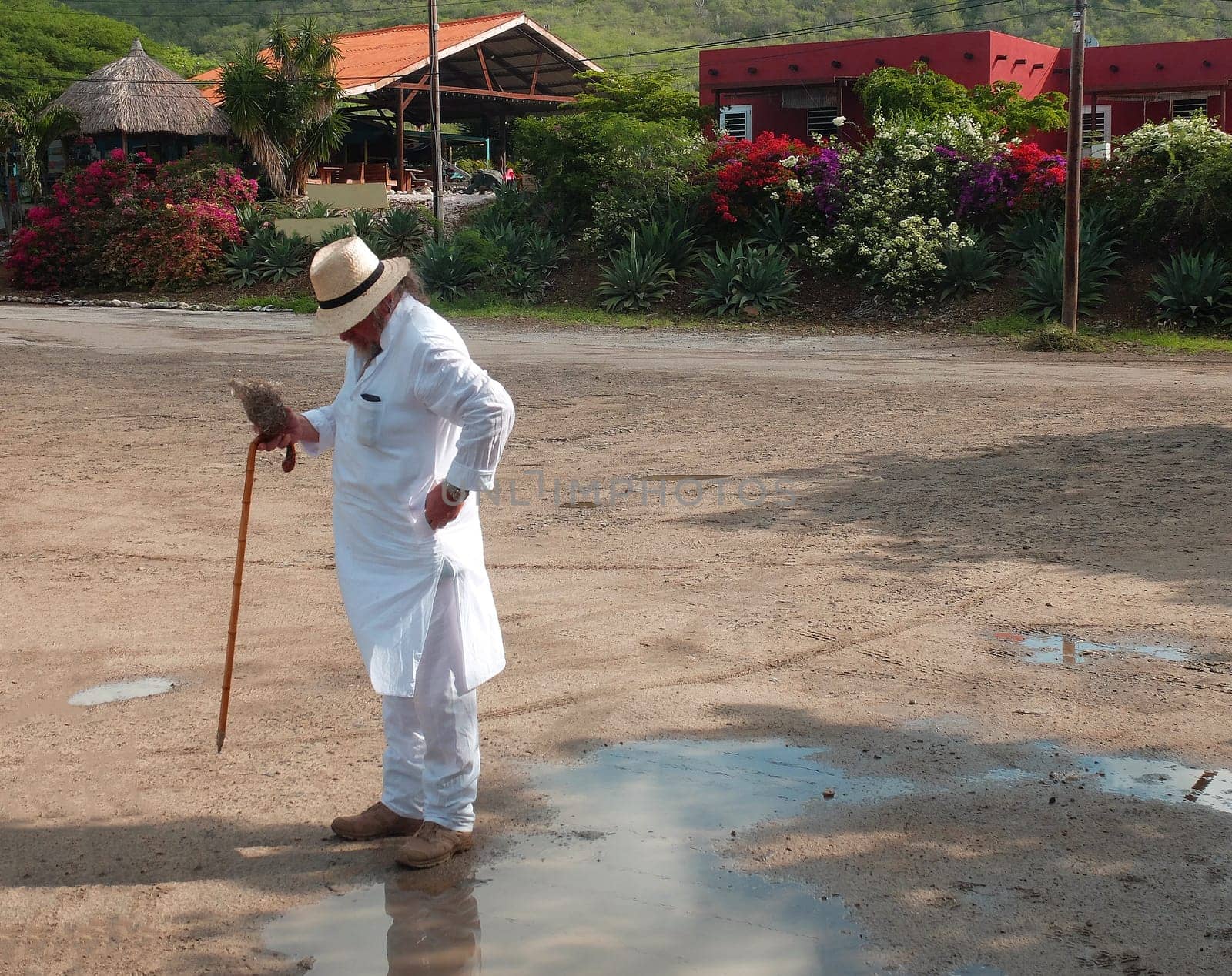 A tourist in white clothes (kurta), a stick and a straw hat standing in front of a row houses. He is checking his pocket. Caribbean vacation.