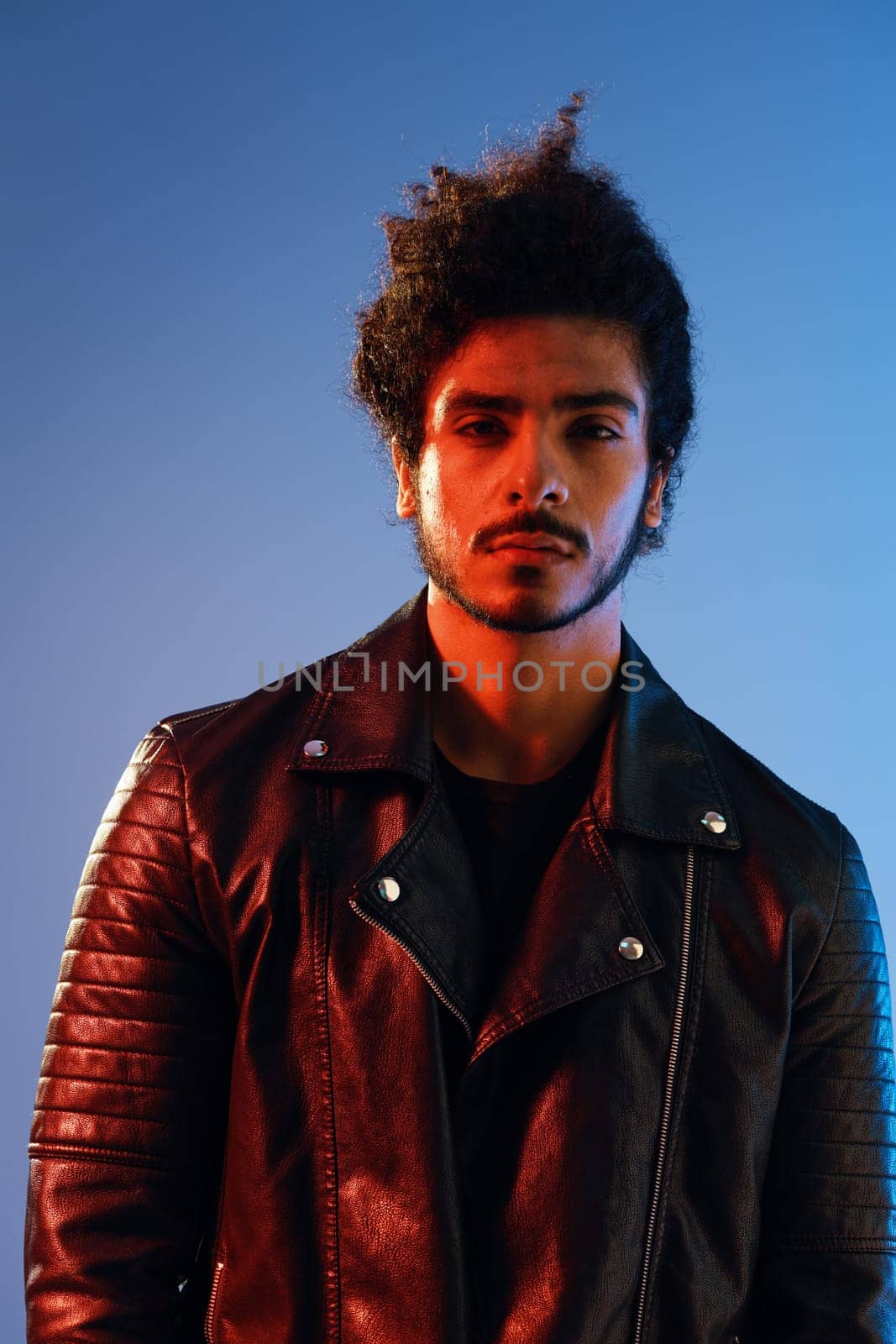 Portrait of a stylish man with curly hair on a blue background multinational, colored light, black leather jacket trend, modern concept. by SHOTPRIME