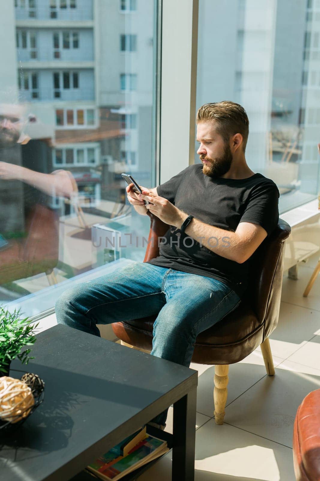 Smiling young adult freelance business man using mobile phone checking social media network feed or message chat sitting on chair. Online digital communication and gadget addiction concept by Satura86