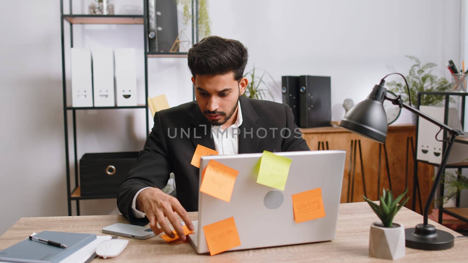 Tired exhausted indian businessman working on laptop at office with many sticker task inscriptions. Freelancer man busy occupation feels sad boredom overworked multitasking. Panic attack, deadline