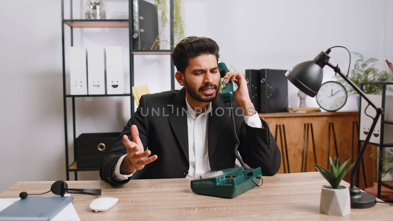 Bearded indian businessman working, having talk with client on retro old-fashioned wired telephone at home office workplace desk. Salesman in formal suit. Remote distant job. Employment occupation