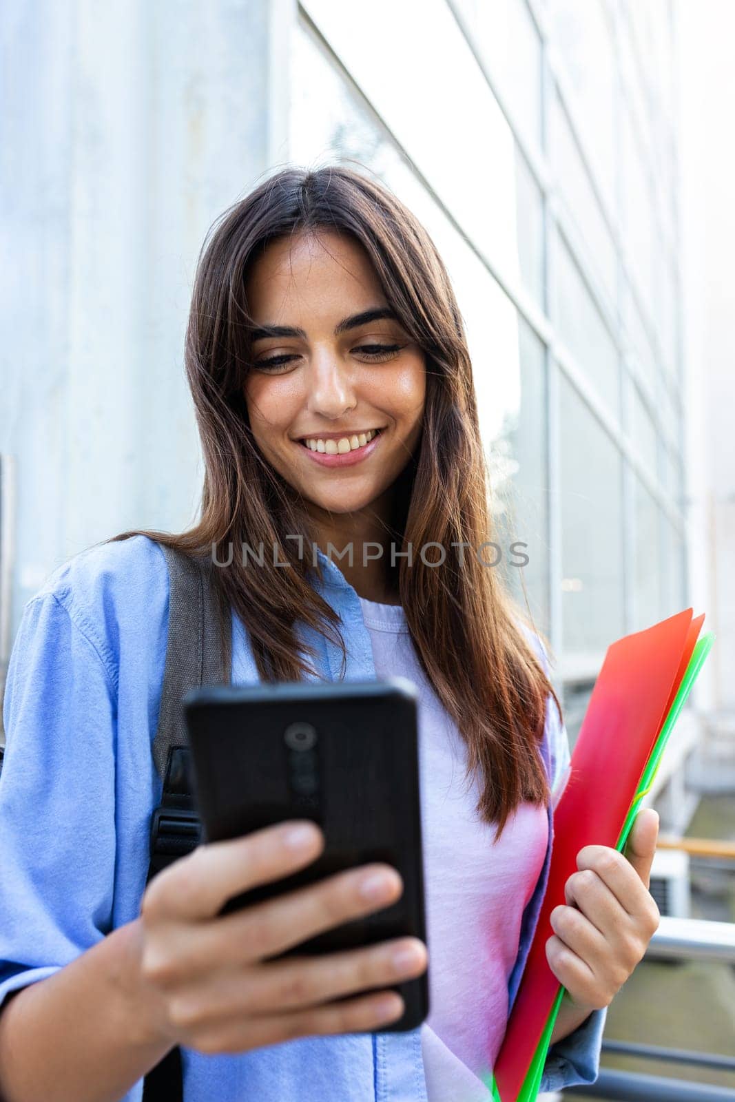 Front view of happy young brunette female university student using smart phone standing outside university building. Vertical image. Education concept.