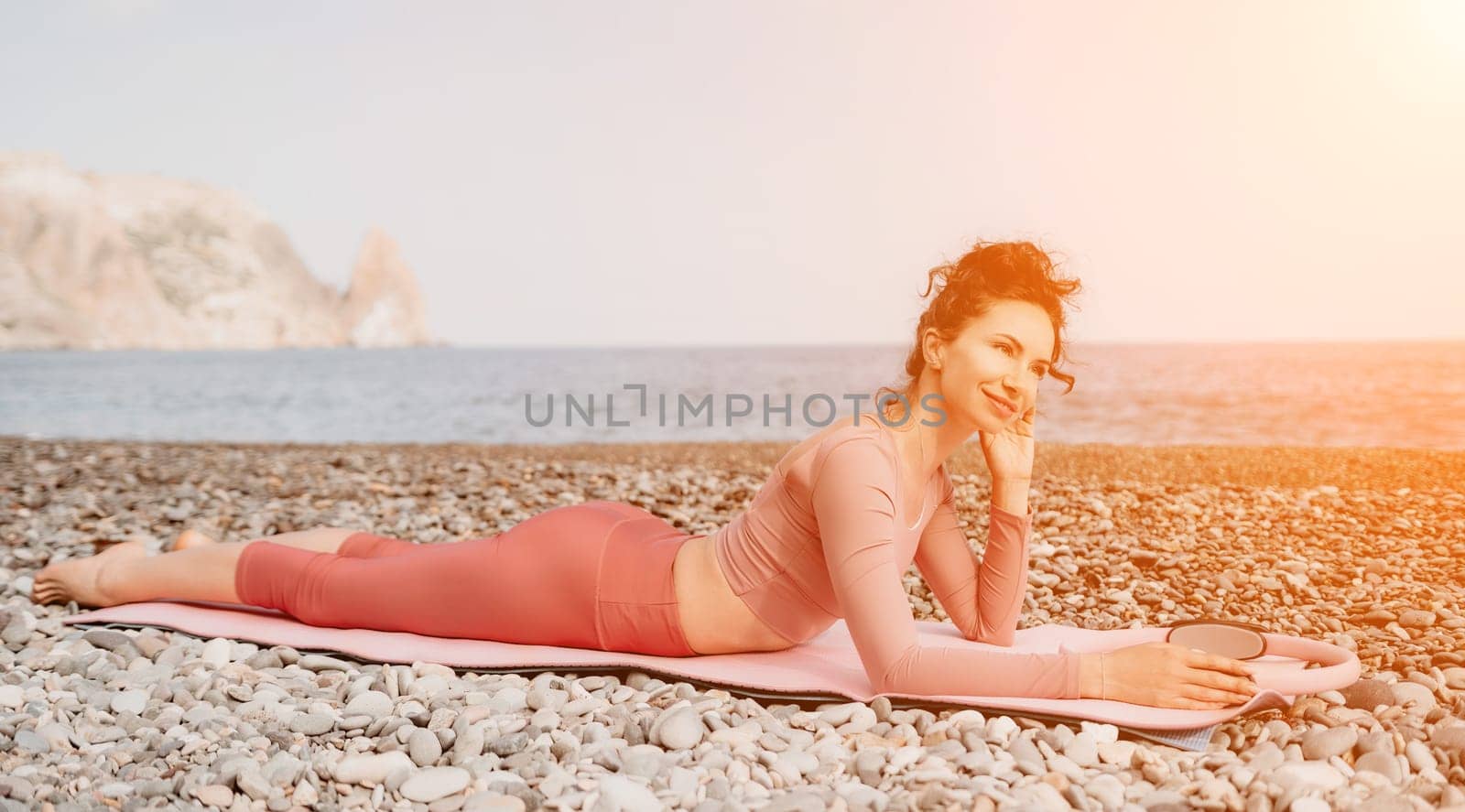 Middle aged well looking woman with black hair doing Pilates with the ring on the yoga mat near the sea on the pebble beach. Female fitness yoga concept. Healthy lifestyle, harmony and meditation.
