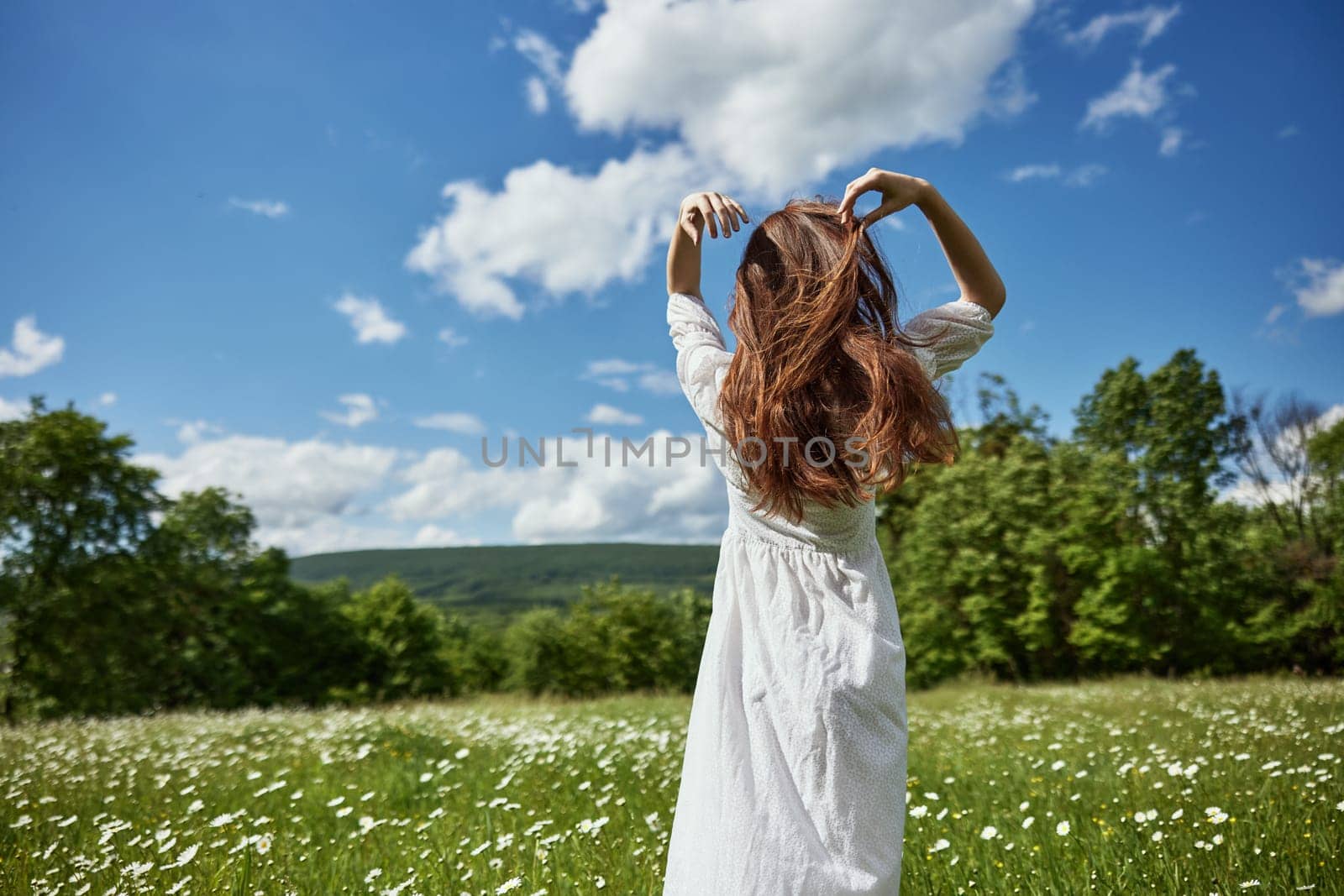 a woman stands in a light dress in a chamomile field and straightens her hair with her hand. High quality photo