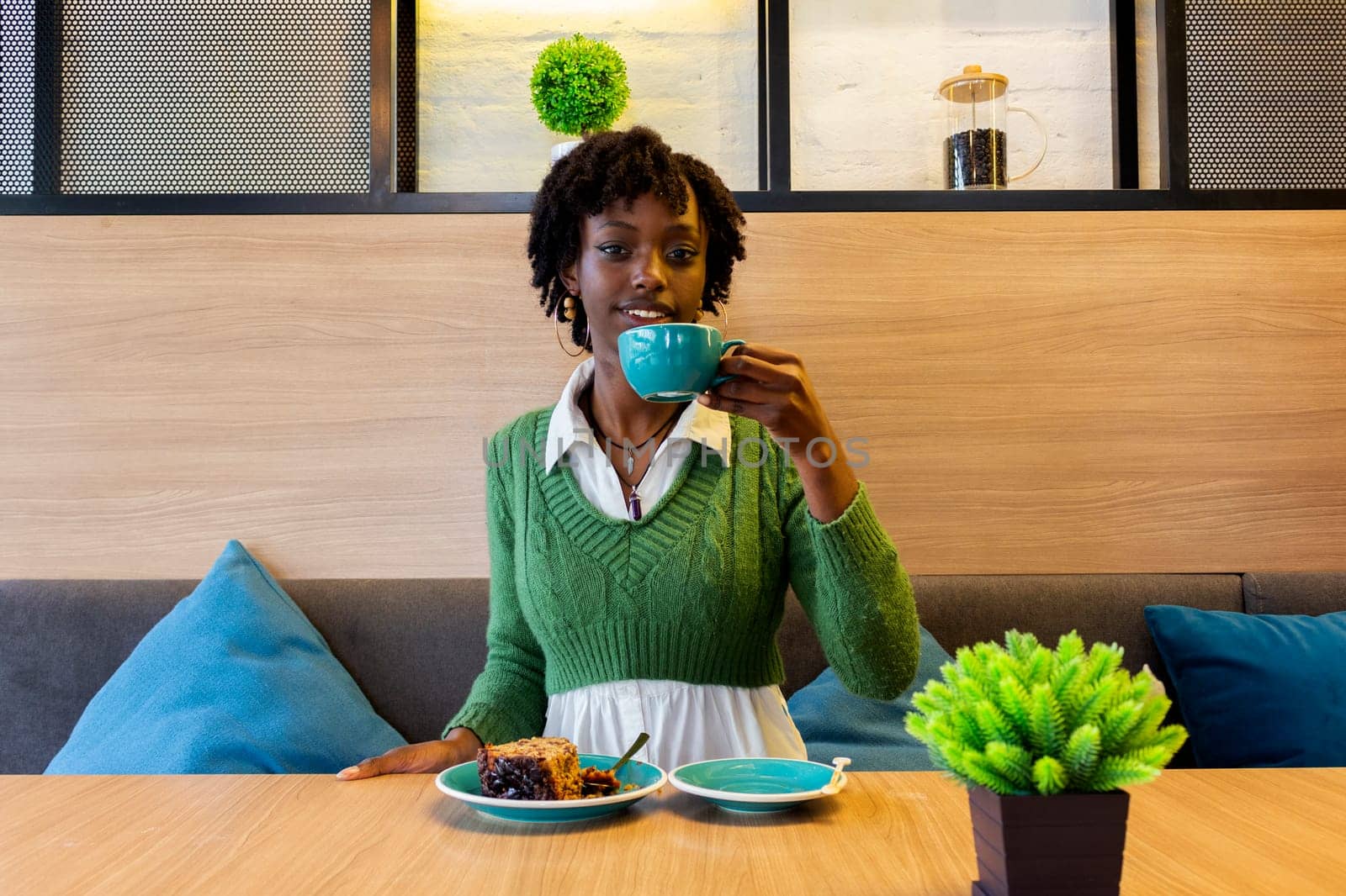 young afro girl sitting in a cafeteria drinking a hot coffee and eating a cake looking at camera. High quality photo