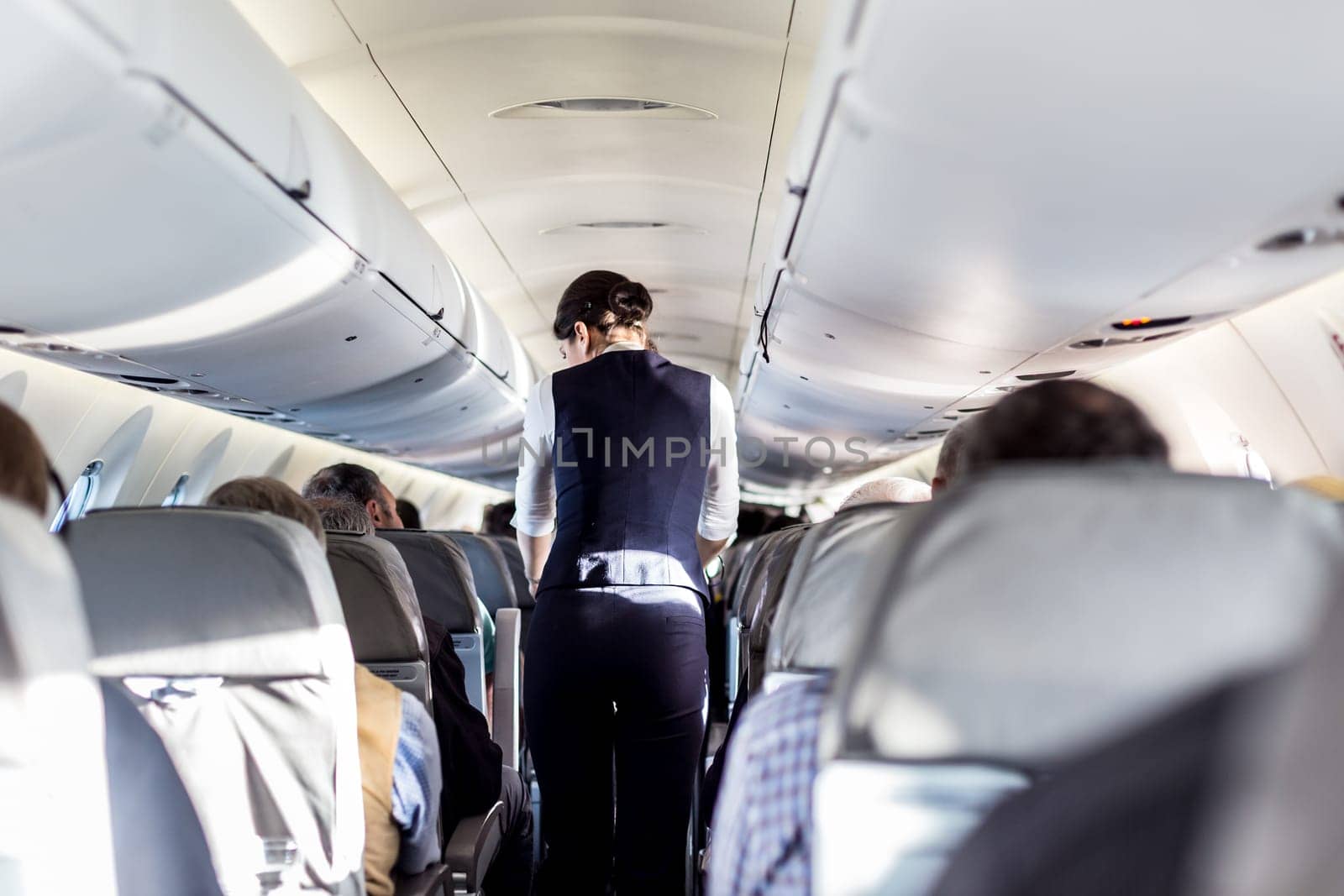 Interior of airplane with passengers on seats and stewardess in uniform walking the aisle, serving people. Commercial economy flight service concept