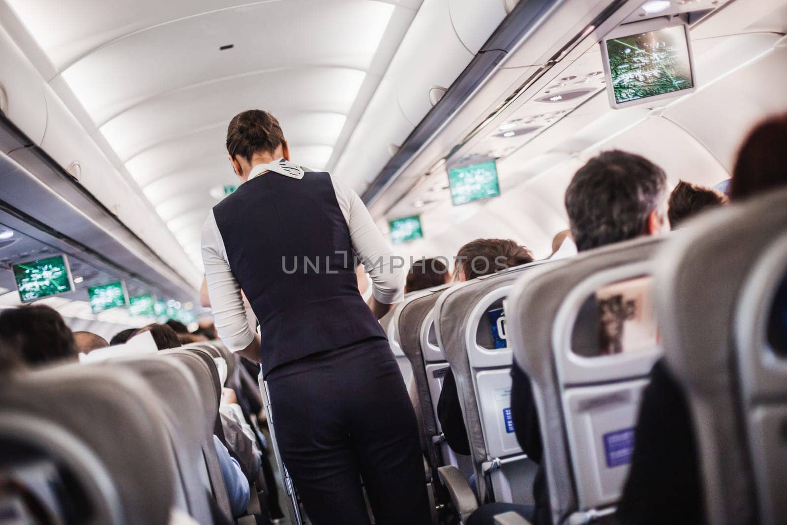 Interior of airplane with passengers on seats and stewardess in uniform walking the aisle, serving people. Commercial economy flight service concept
