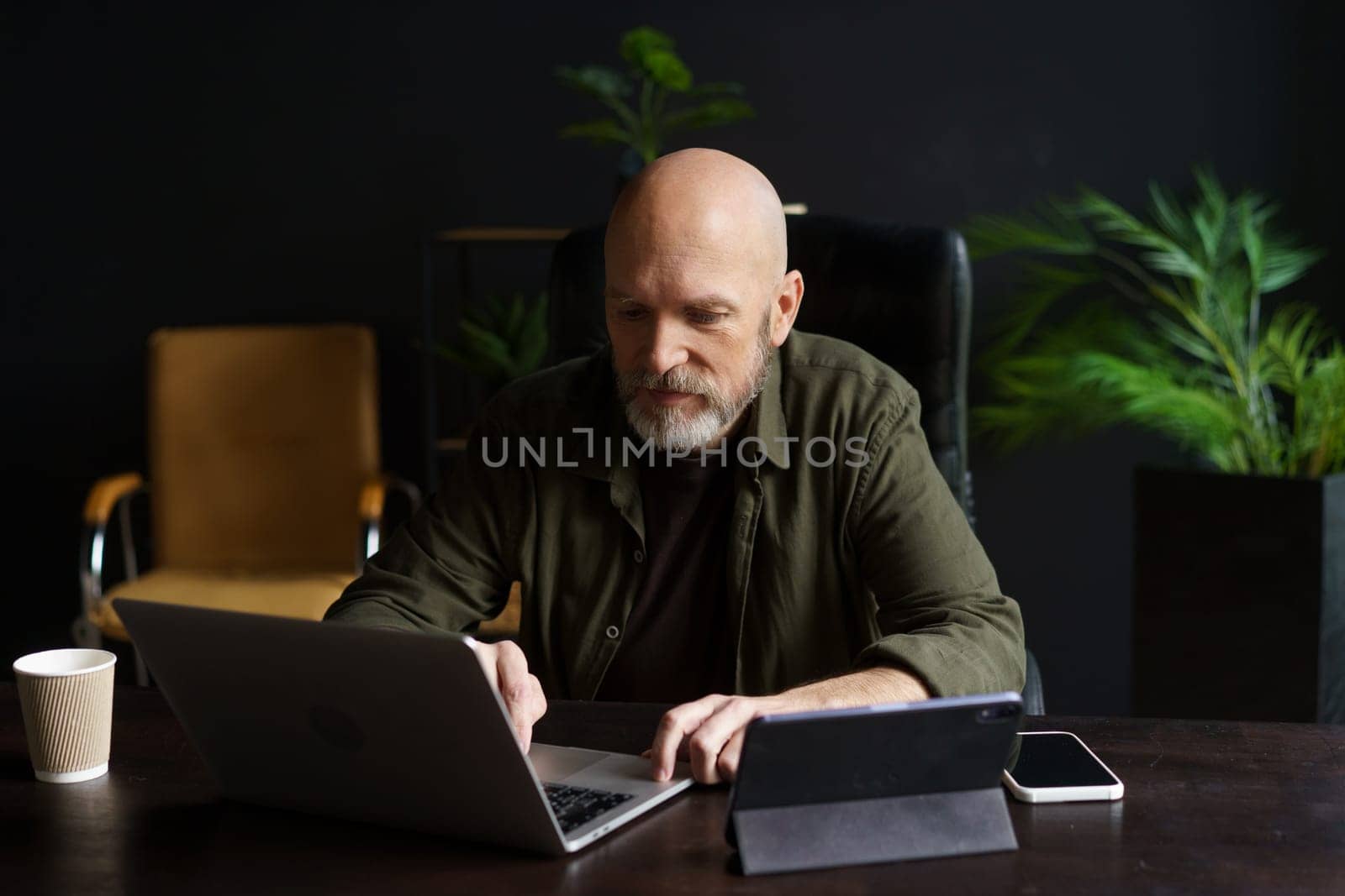 Man diligently working on his laptop from the comfort of his home. With a dedicated workspace and a focused expression, he embraces the concept of online work and remote productivity. by LipikStockMedia
