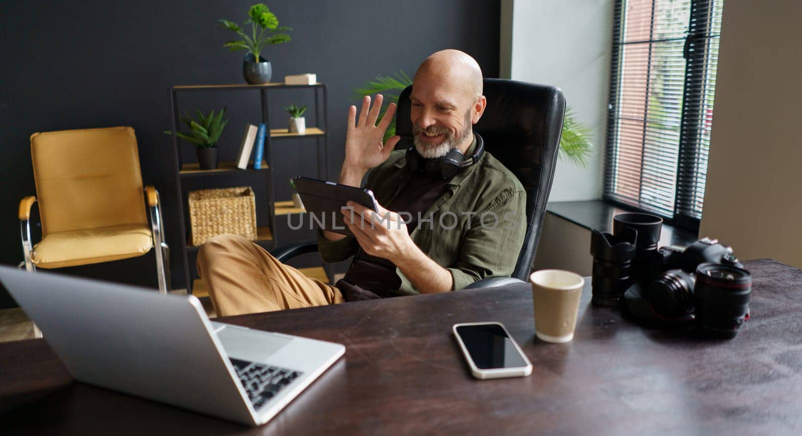 Photographer has online call with client. He holds Tablet PC and welcoming interlocutor. Desk in front equipped with laptop, mobile phone, and photo camera with lenses, indicating tools of business. High quality photo