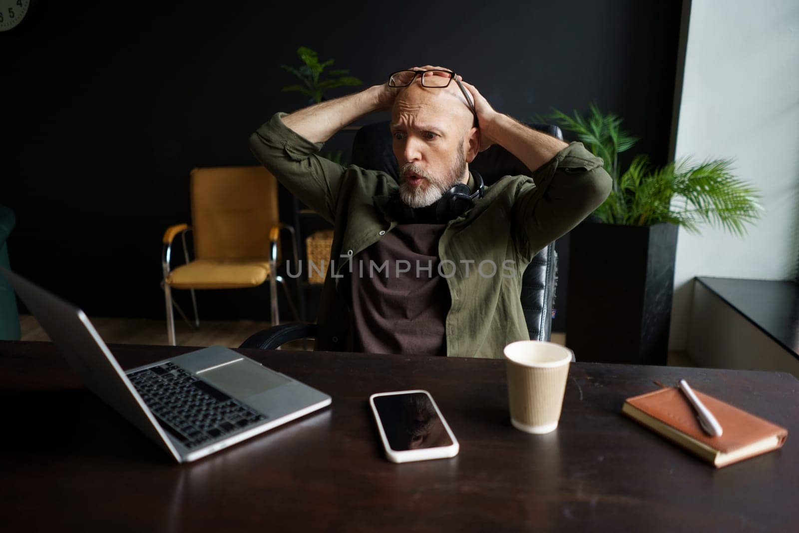 Man sitting in chair in loft office with unpleasant surprise. His face shows mixture of shock and disbelief, and he screaming with hands raised in air. Desk holding a laptop and other office supplies. High quality photo