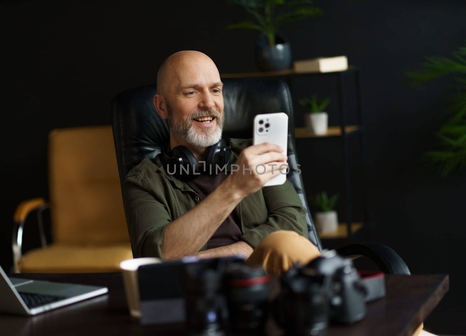 Mid aged man with bald head and silver beard engrossed in reading news on mobile phone. With laptop placed on desk in background, image captures essence of connected and digitally-oriented lifestyle. . High quality photo