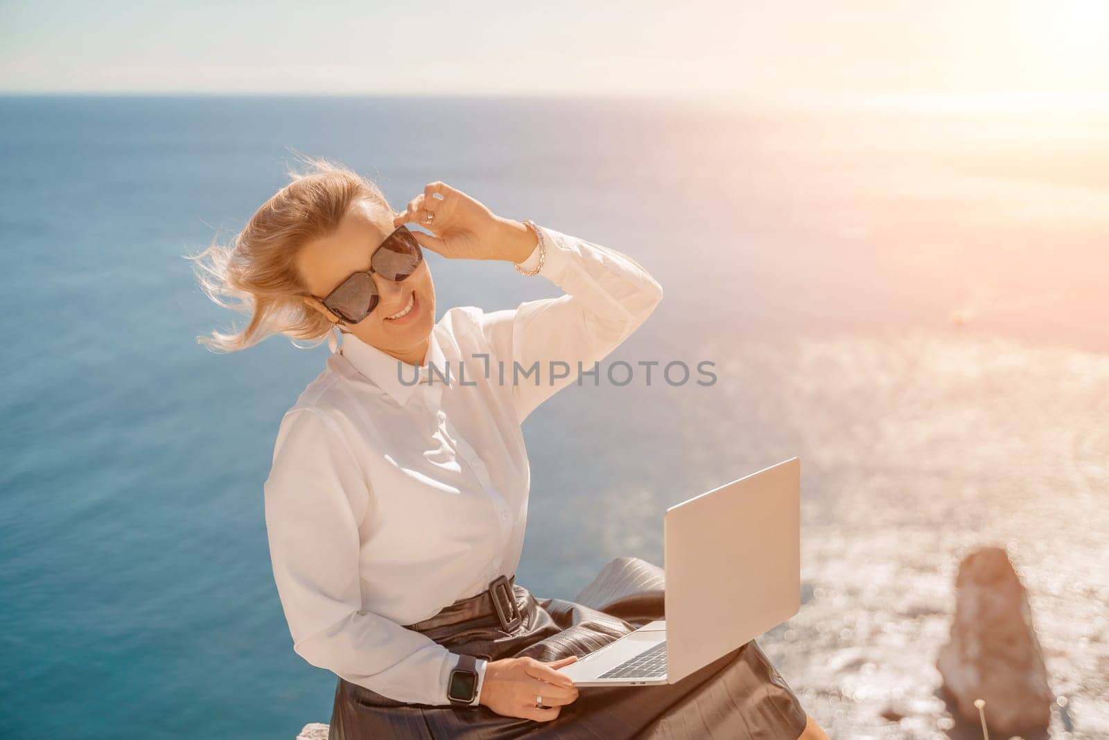 Business woman on nature in white shirt and black skirt. She works with an iPad in the open air with a beautiful view of the sea. The concept of remote work. by Matiunina