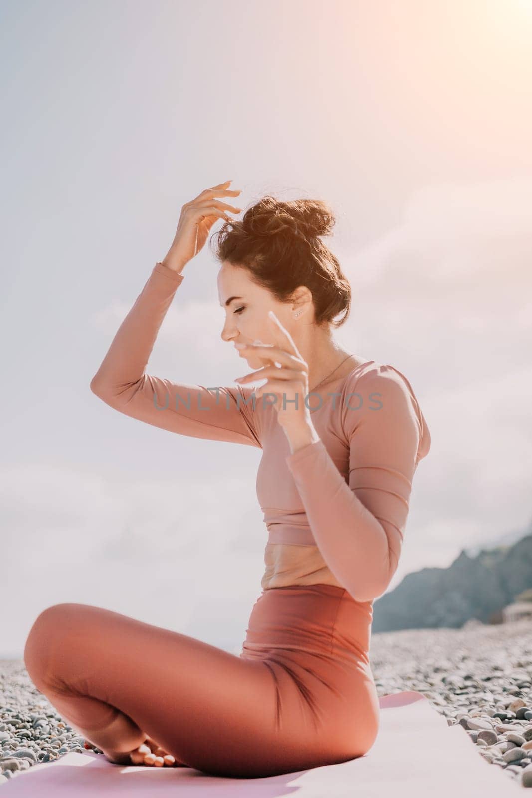 Young woman with long hair in white swimsuit and boho style braclets practicing outdoors on yoga mat by the sea on a sunset. Women's yoga fitness routine. Healthy lifestyle, harmony and meditation