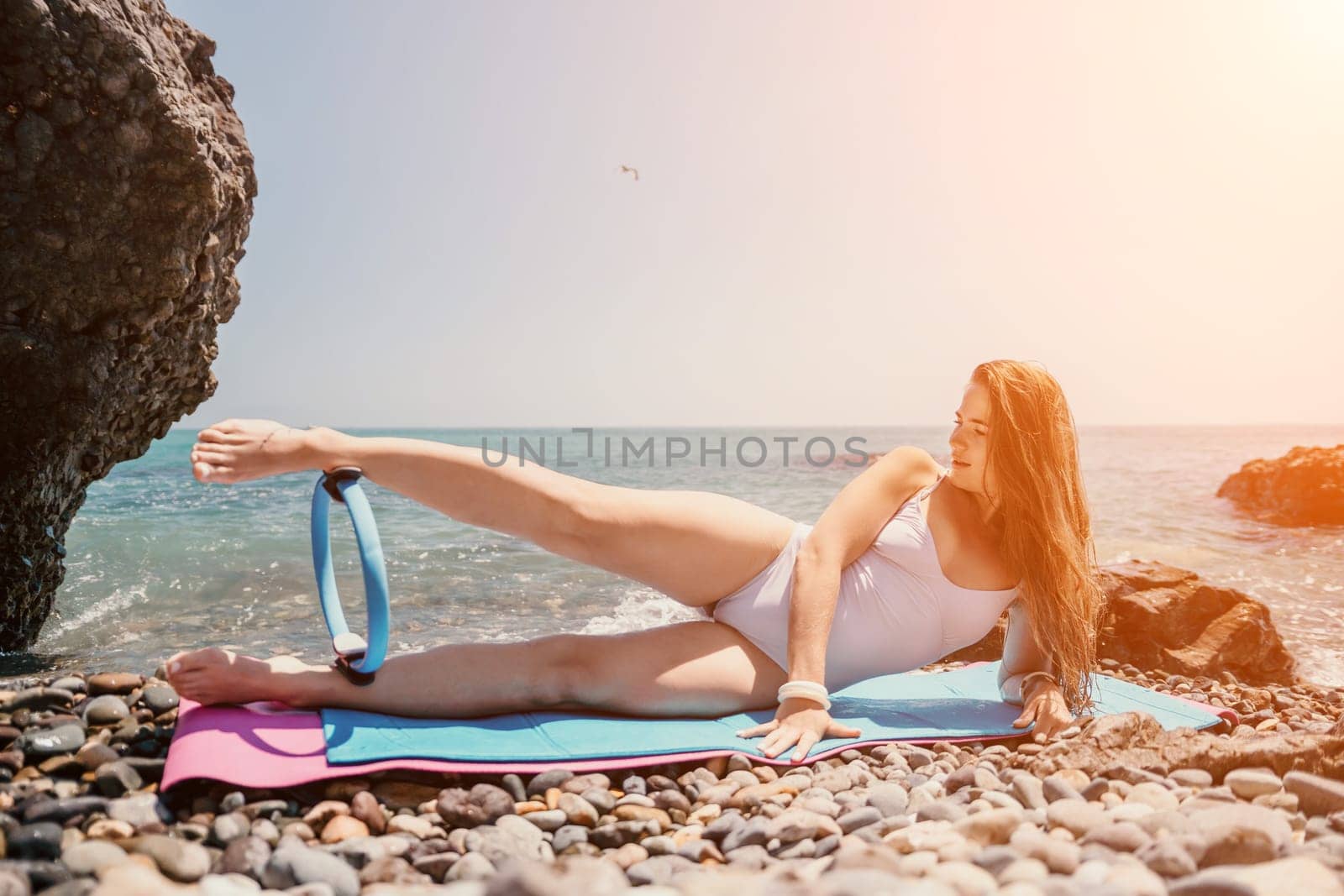 Young woman with black hair, fitness instructor in pink sports leggings and tops, doing pilates on yoga mat with magic pilates ring by the sea on the beach. Female fitness daily yoga concept