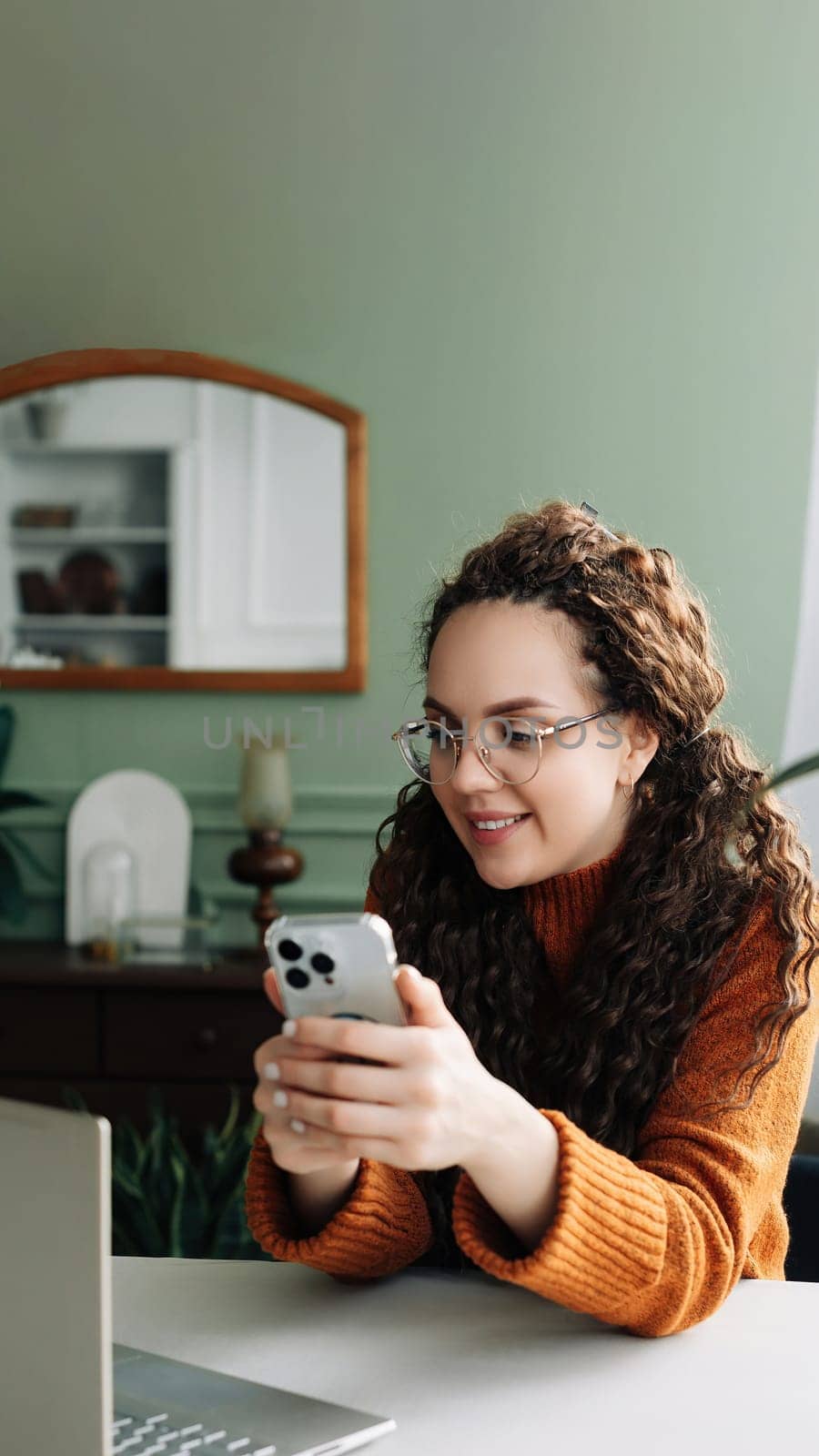 Young happy smiling pretty woman sitting at table holding smartphone using cellphone modern technology, looking at mobile phone while remote working or learning, texting messages at home