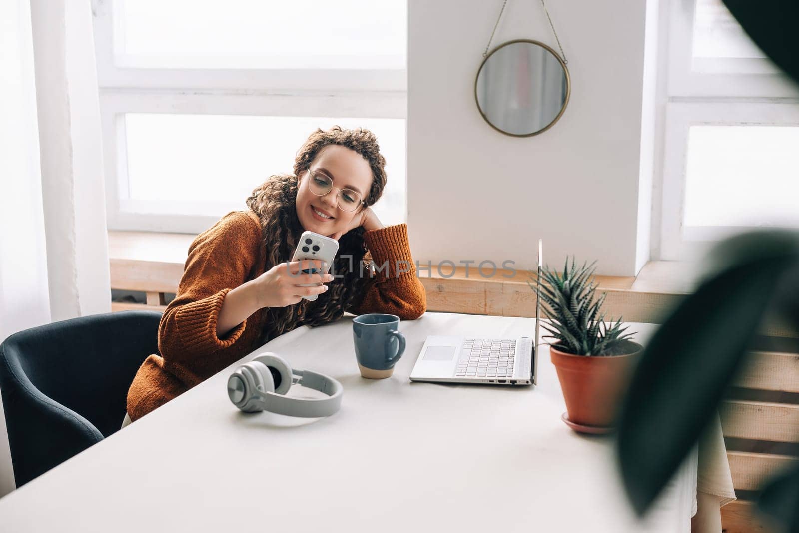 Young happy smiling pretty woman sitting on chair holding smartphone using cell phone mobile technology, looking at mobile phone while remote working or learning, texting messages at home office.