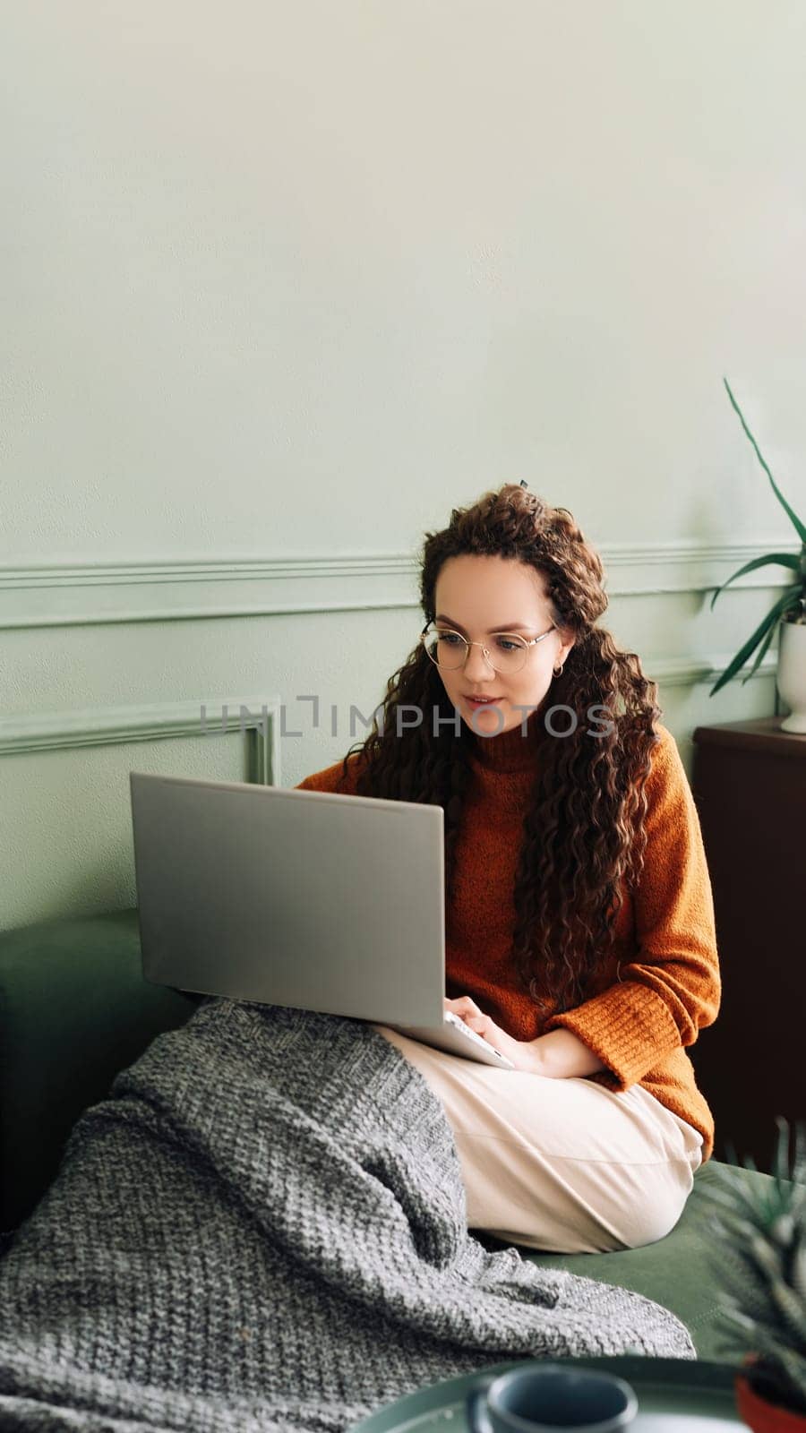Relaxed young woman sitting on sofa holding mobile phone using cellphone and laptop technology doing online ecommerce shopping, texting messages relaxing on couch in cozy living room home interior