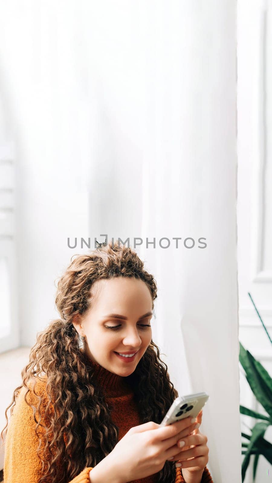 Young woman browsing online on cellphone while sitting comfortably. Happy woman shopping on smartphone while relaxing on couch. Smiling lady using smartphone for social media and e-commerce.