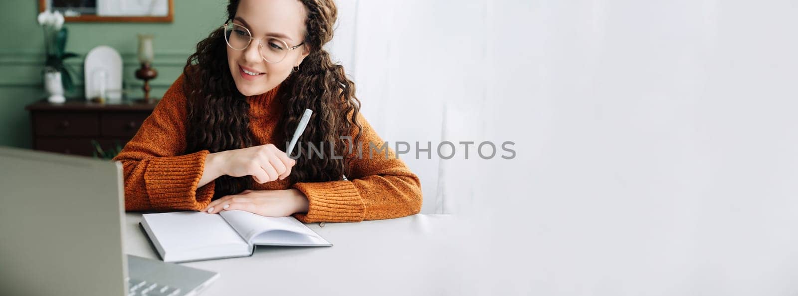 Young smiling pretty woman student using laptop elearning or remote working at home office using laptop computer watching webinar, learning web course, studying online sitting at table, writing notes