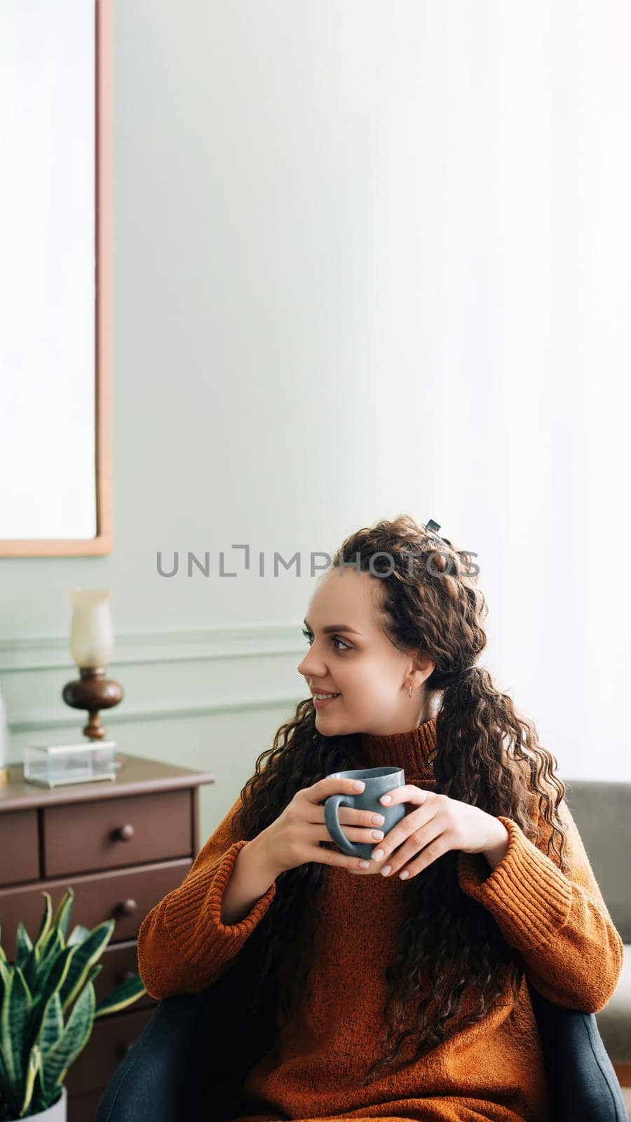 Dreamy young woman savoring warm coffee in cozy home interior. Beautiful lady enjoying a hot drink while relaxing at home. Serene woman with warm mug, lost in thought on cozy winter day.