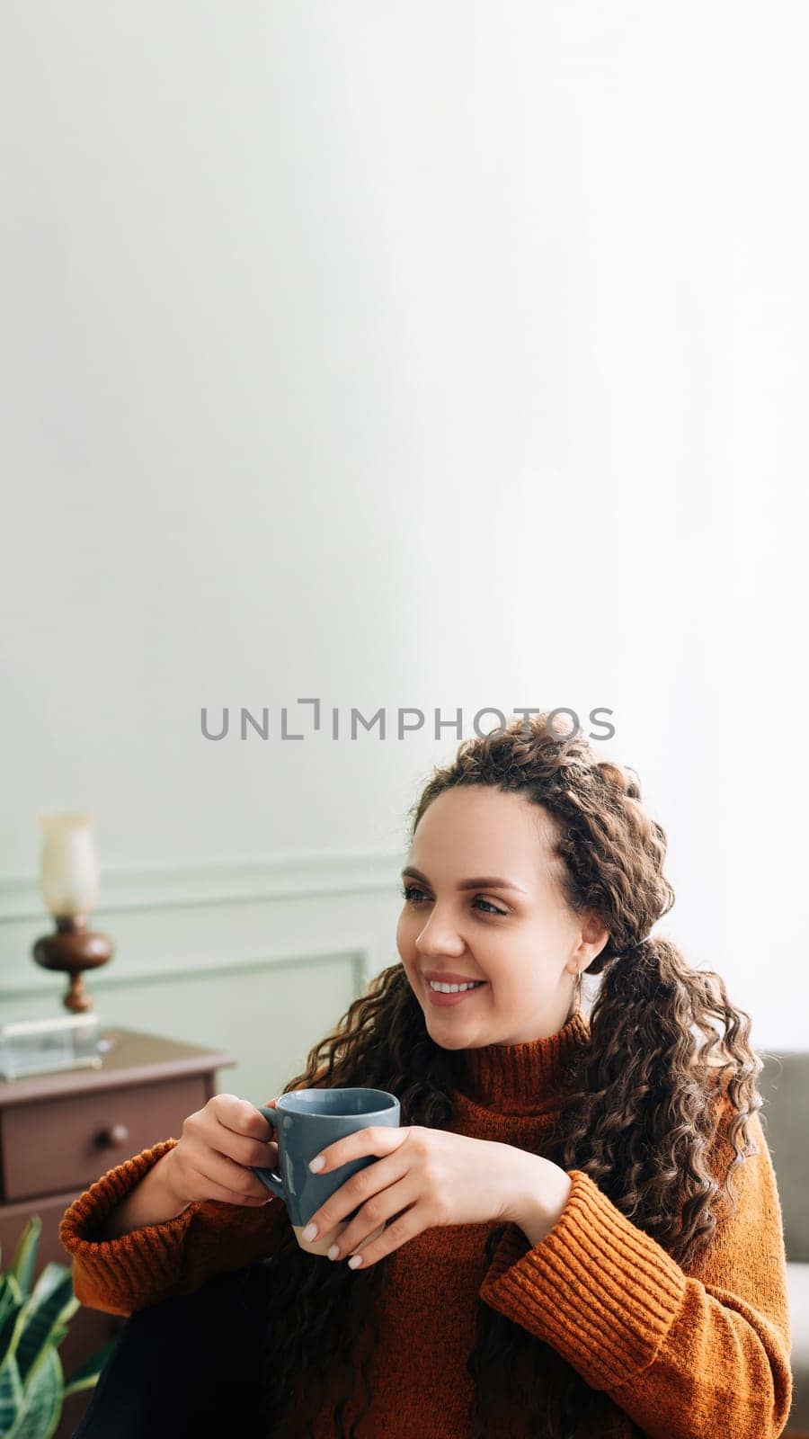 Cozy Winter Mornings: Happy Woman Enjoying Warm Drink and Daydreaming at Home. Warmth and Relaxation: Young Woman Holding a Cup of Coffee and Daydreaming at Home