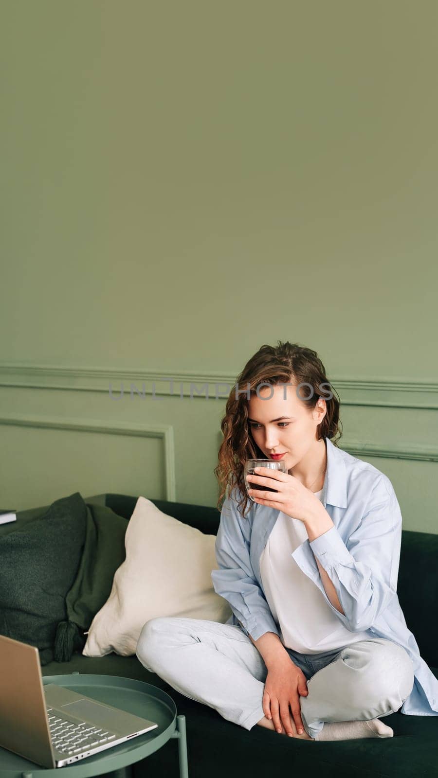 Adorable female drinking hot tea sitting on a sofa near a big window and watching webinars and online tutorials on a laptop using a wireless connection, studying from home.
