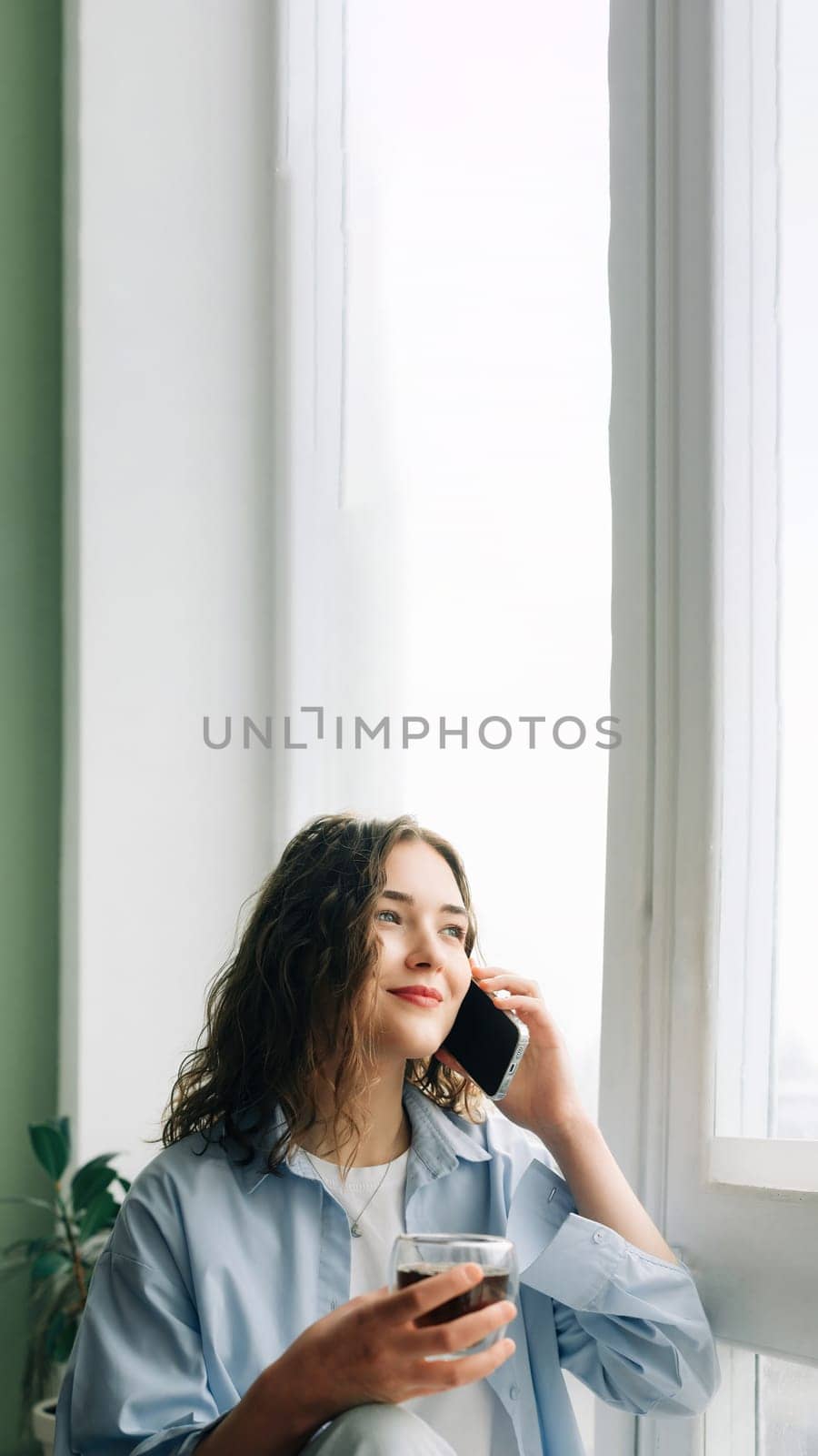 Cheerful young woman in casual attire chatting and flirting with her boyfriend on the phone, sitting on a windowsill with a cup of black tea. by ViShark