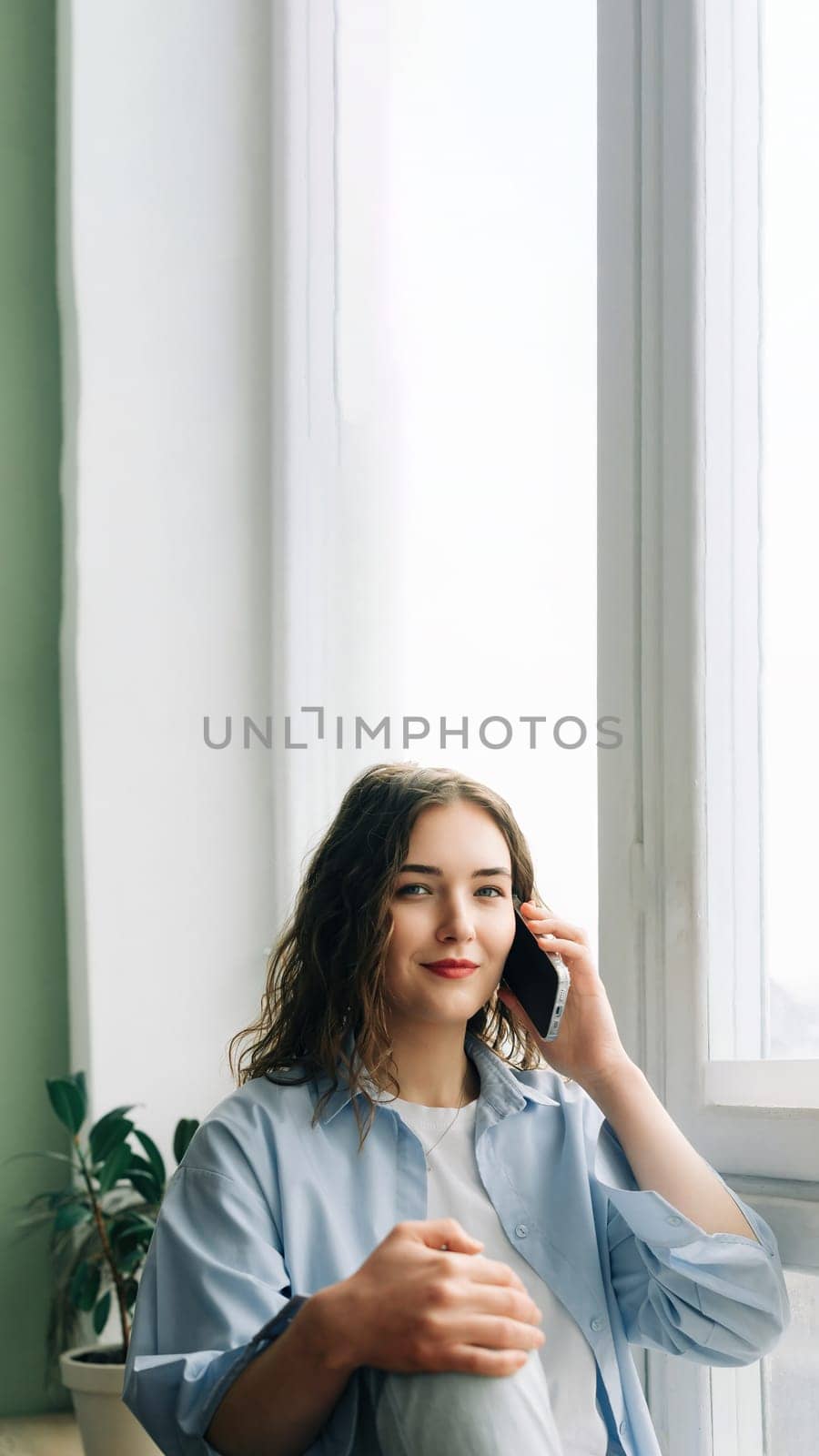 A busy but happy mom enjoying a phone call with her best friend while kids play in the background. Casual indoor lifestyle portrait of a charming 30s woman sitting on windowsill by ViShark