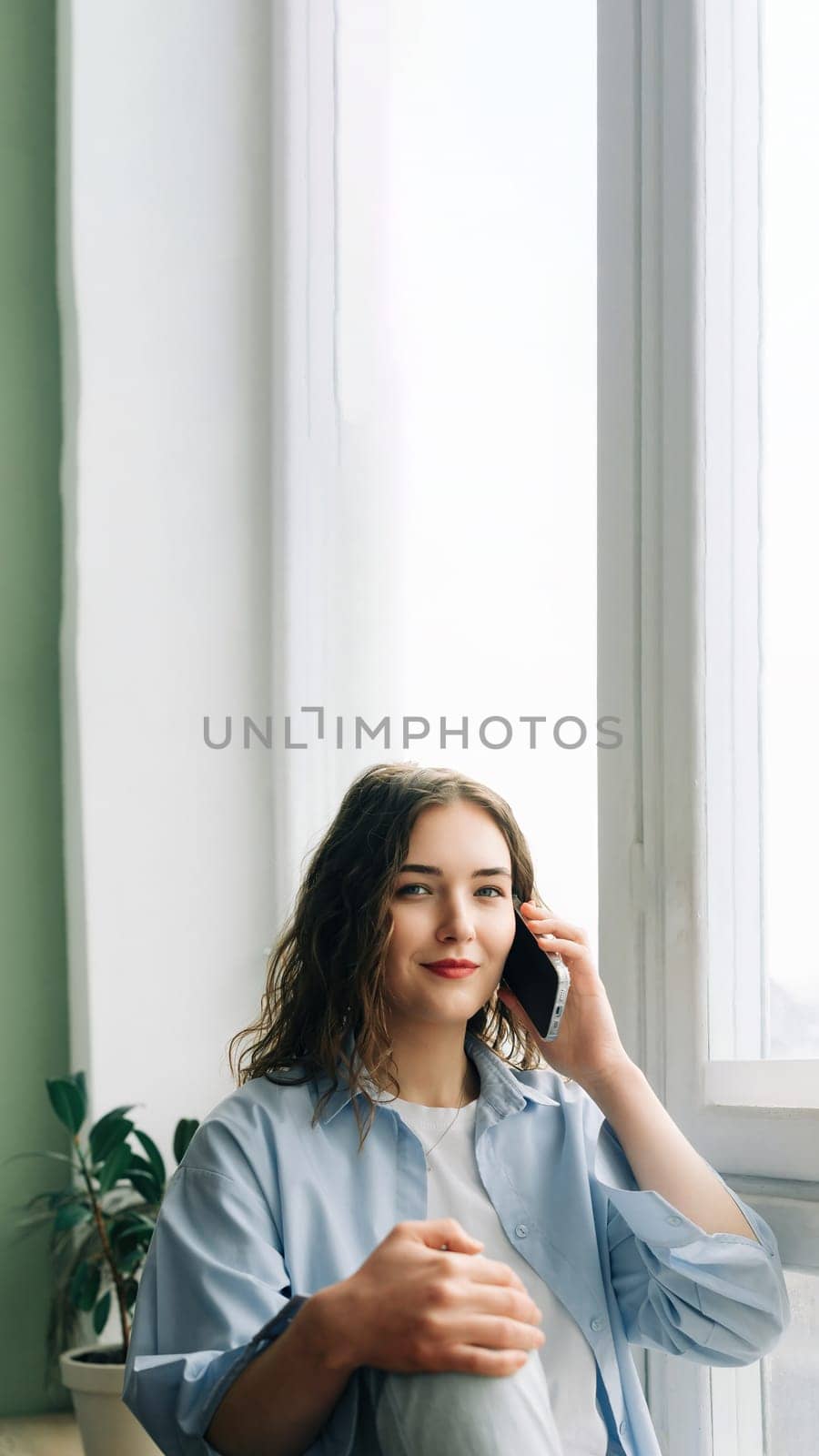 A busy but happy mom enjoying a phone call with her best friend while kids play in the background. Casual indoor lifestyle portrait of a charming 30s woman sitting on windowsill.