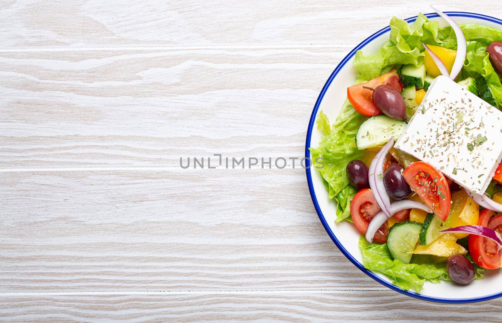 Traditional Greek Salad with Feta Cheese, Tomatoes, Bell Pepper, Cucumbers, Olives, Herbs in white ceramic bowl on White rustic wooden table background from above, Cuisine of Greece. Copy space