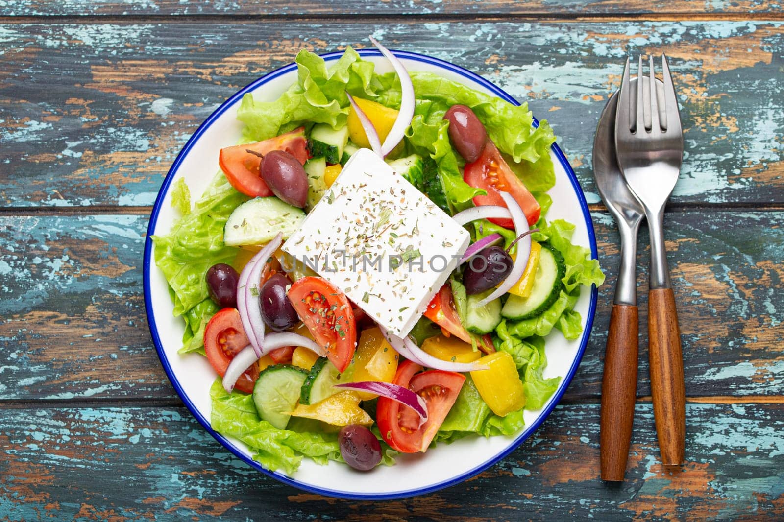 Traditional Greek Salad with Feta Cheese, Tomatoes, Bell Pepper, Cucumbers, Olives, Herbs in white ceramic bowl on blue rustic wooden table background from above, Cuisine of Greece