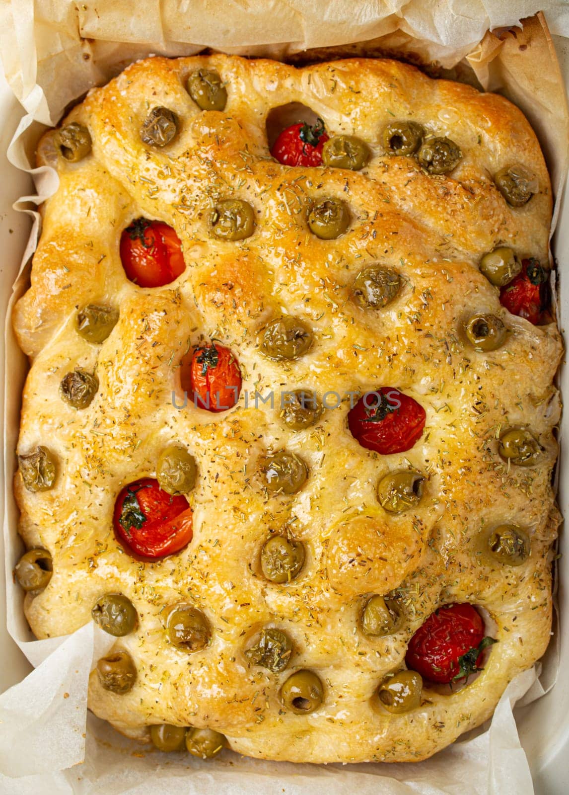 Overhead of Traditional Italian Homemade Flat Bread Focaccia with Green Olives, Olive Oil, Cherry Tomatoes and Rosemary in Baking Tray on Rustic Dark Blue Concrete Background