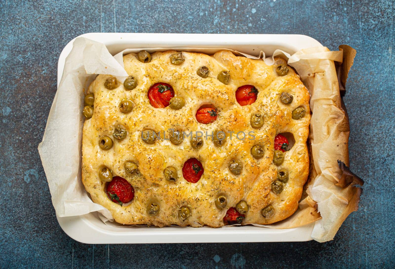 Overhead of Traditional Italian Homemade Flat Bread Focaccia with Green Olives, Olive Oil, Cherry Tomatoes and Rosemary in Baking Tray on Rustic Dark Blue Concrete Background