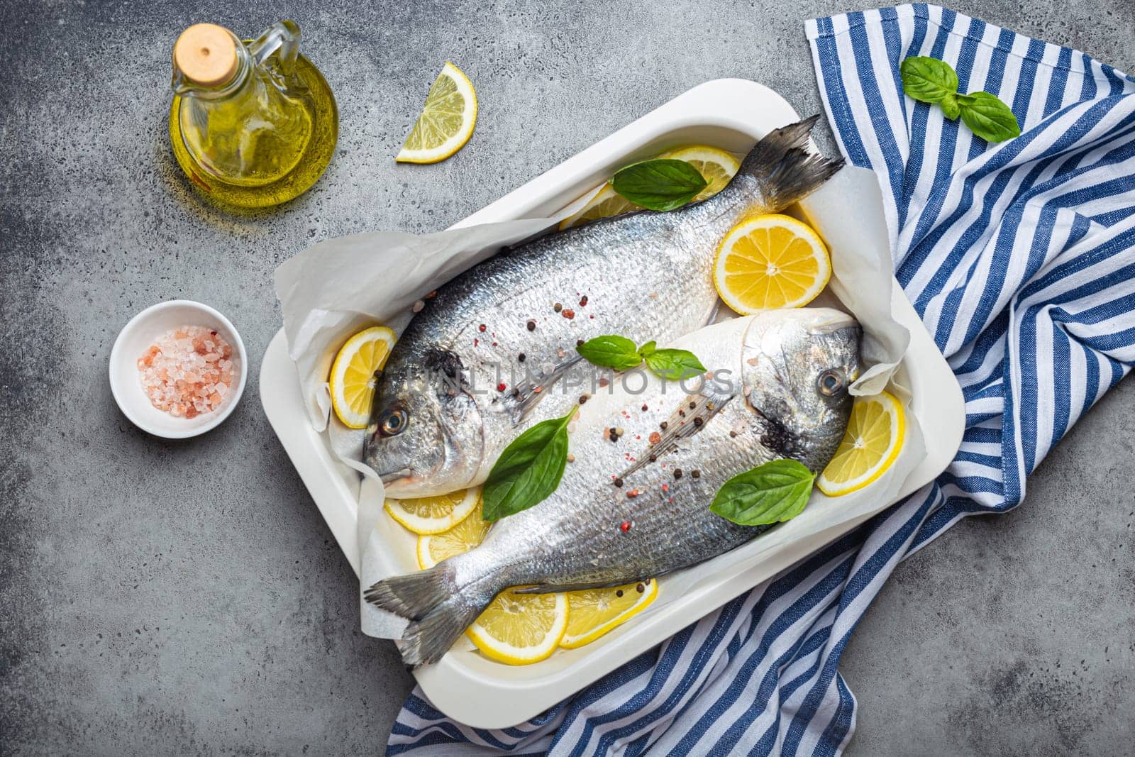 Raw fish dorado in casserole dish with ingredients lemon, fresh basil, bottle of olive oil on wooden cutting board with knife on rustic stone background top view, cooking healthy fish dorado concept