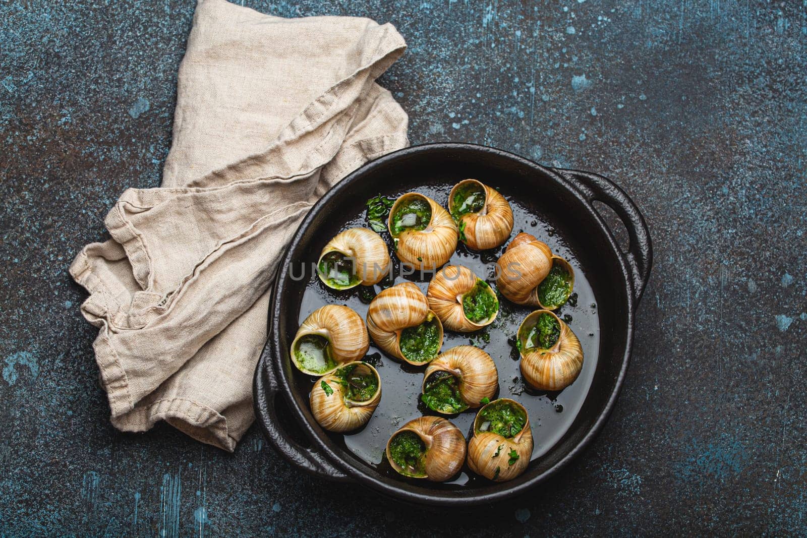 Escargots de Bourgogne Cooked Snails with Garlic Butter and Parsley in black cast iron pan on rustic stone background top view, traditional French Delicacy .
