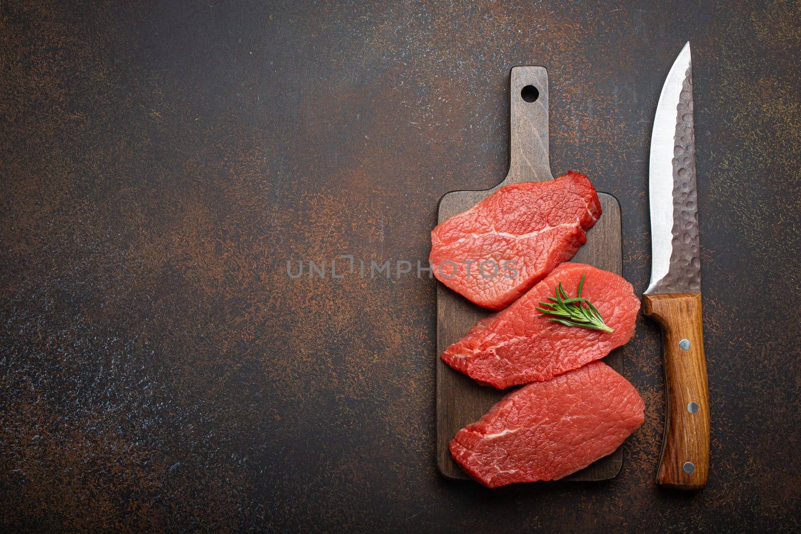 Three raw uncooked meat beef top sirloin steaks on wooden cutting board with rosemary on wooden cutting board with knife on dark rustic background from above, preparing meal with meat, copy space