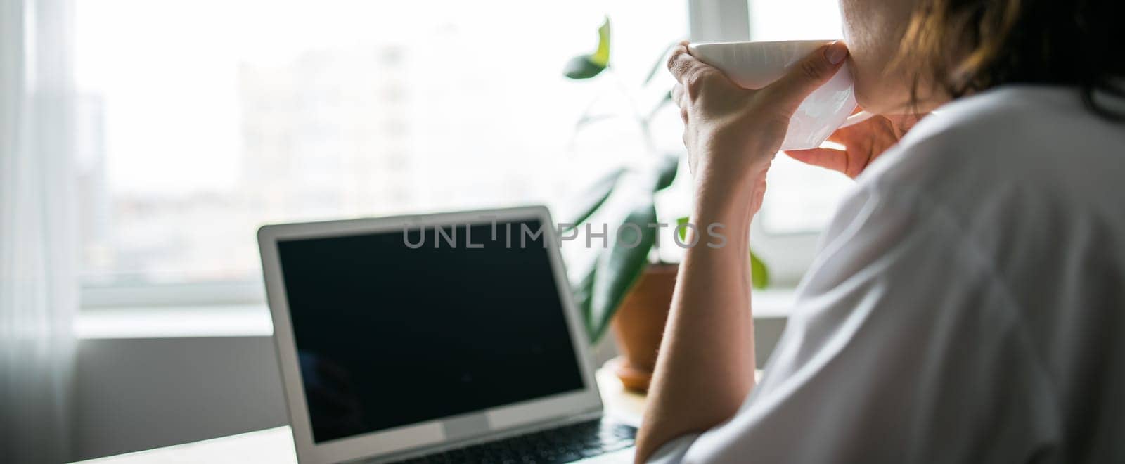 Woman drinks coffee sitting at laptop keyboard on window background. Cup of hot drink in female hand and break during work concept