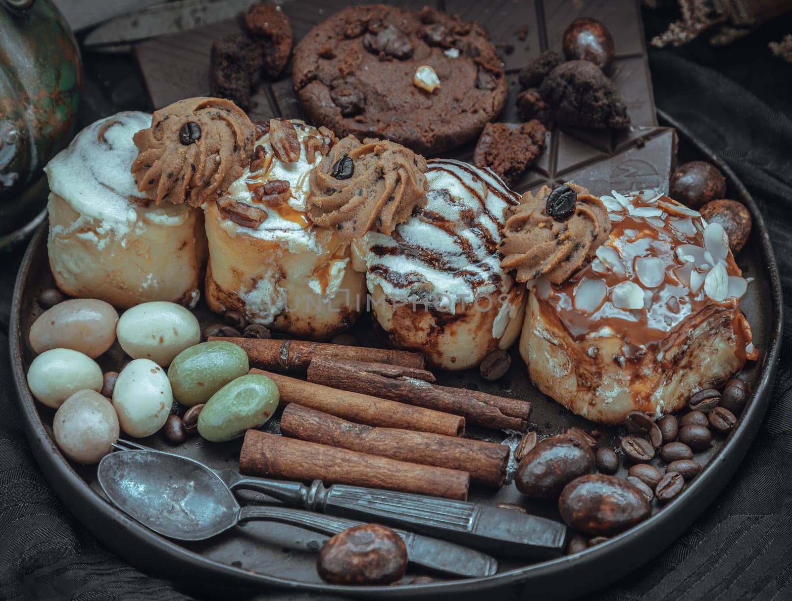 Assorted Cinnamon Rolls, Dark chocolate bars, cookies, pieces and balls served with black iron cutlery in black ceramic plate on dark background. Selective Focus.