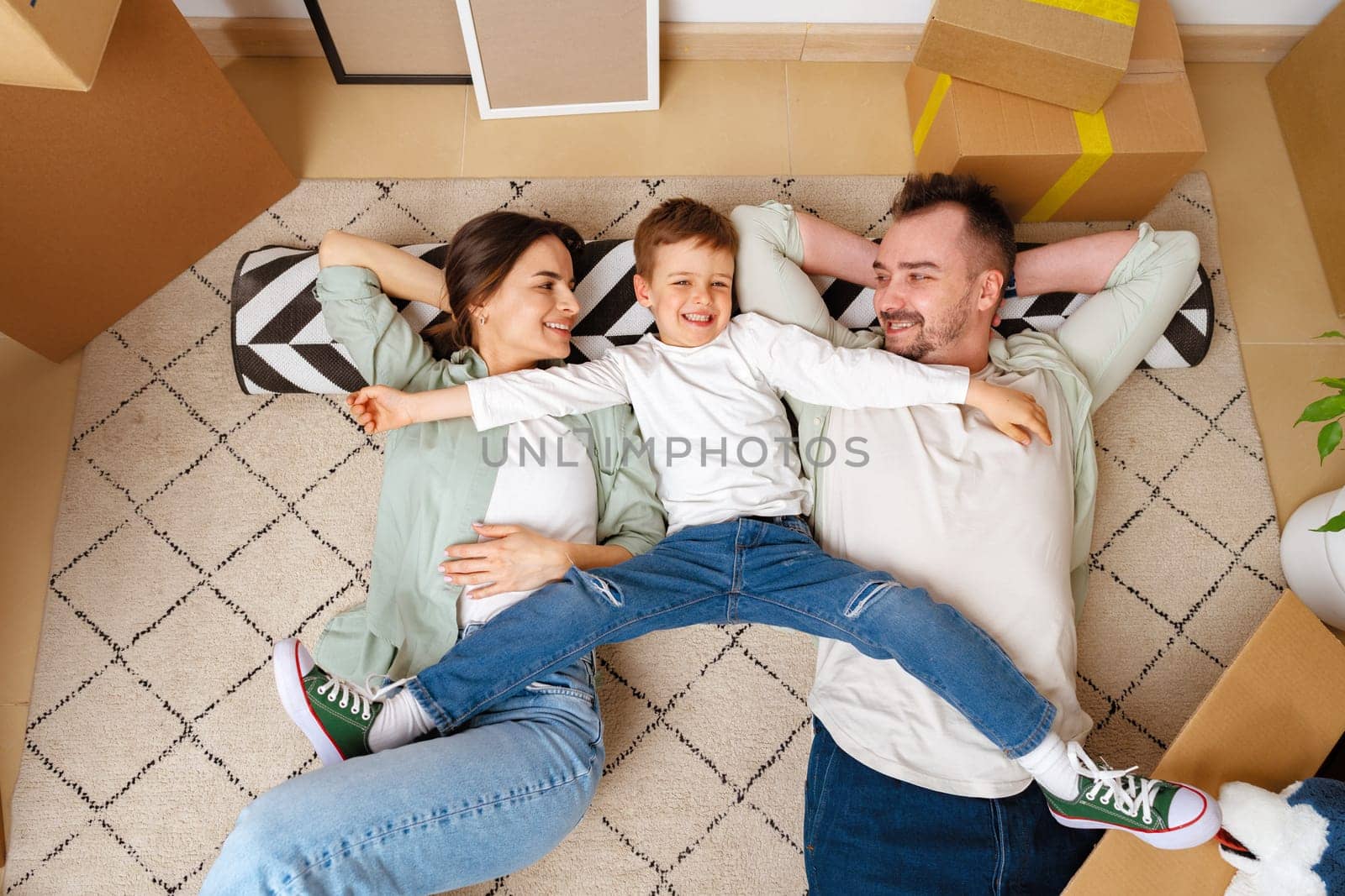 Happy family lying on the floor in new home with cordboard boxes around by Fabrikasimf