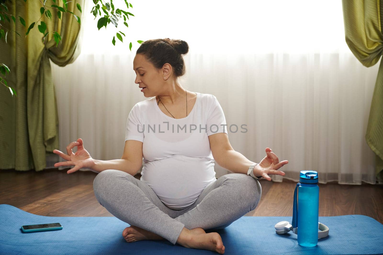 Pregnant woman watching online video lesson with pregnancy yoga exercises, on mobile app in her smartphone, sitting in lotus position on a blue fitness mat, practicing yoga, stretching and meditation