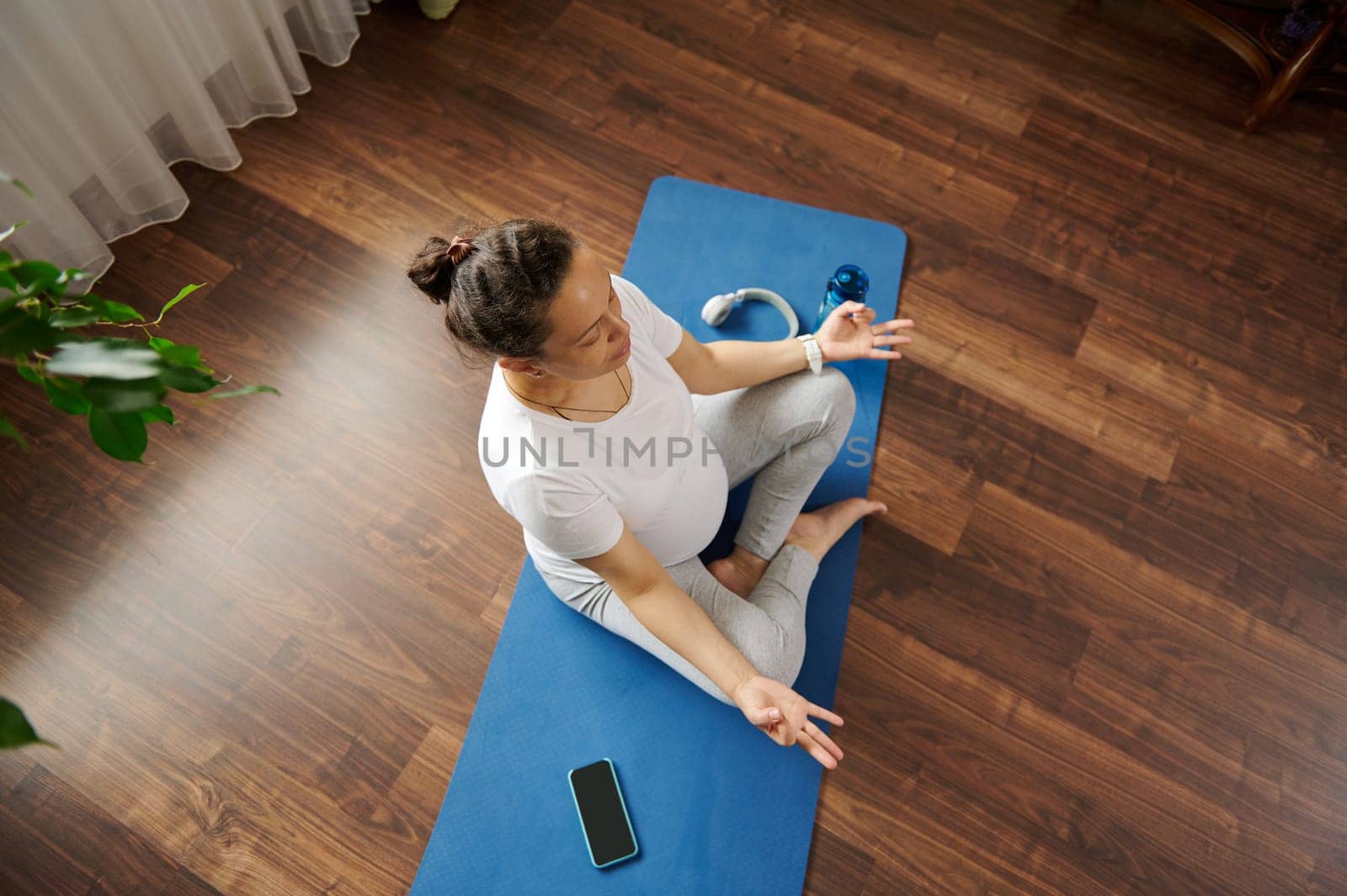 Top view peaceful pregnant woman yogi holding fingers in mudra gesture, meditating with her eyes closed, feeling calm, positive and relaxed. Yoga practice and meditation for healthy active pregnancy