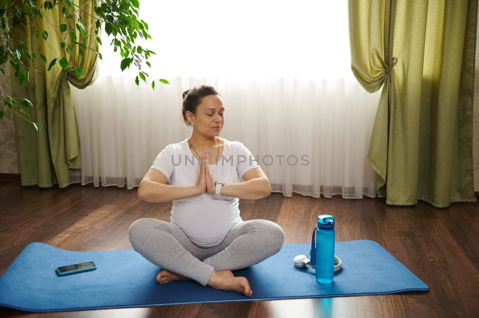 Delightful ethnic pregnant woman with closed eyes, meditating on lotus pose on yoga mat, with hands palms together. Happy gravid female enjoying healthy lifestyle and positive emotions in pregnancy