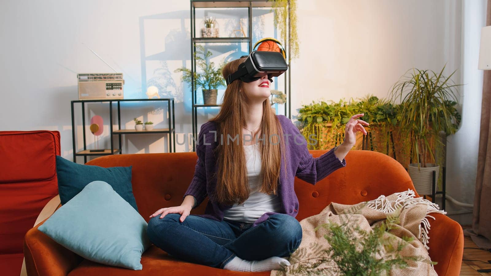 Portrait of woman using virtual reality futuristic technology VR app headset helmet to play simulation 3D 360 video game, watching film movie at modern home apartment. Girl in goggles sitting on couch