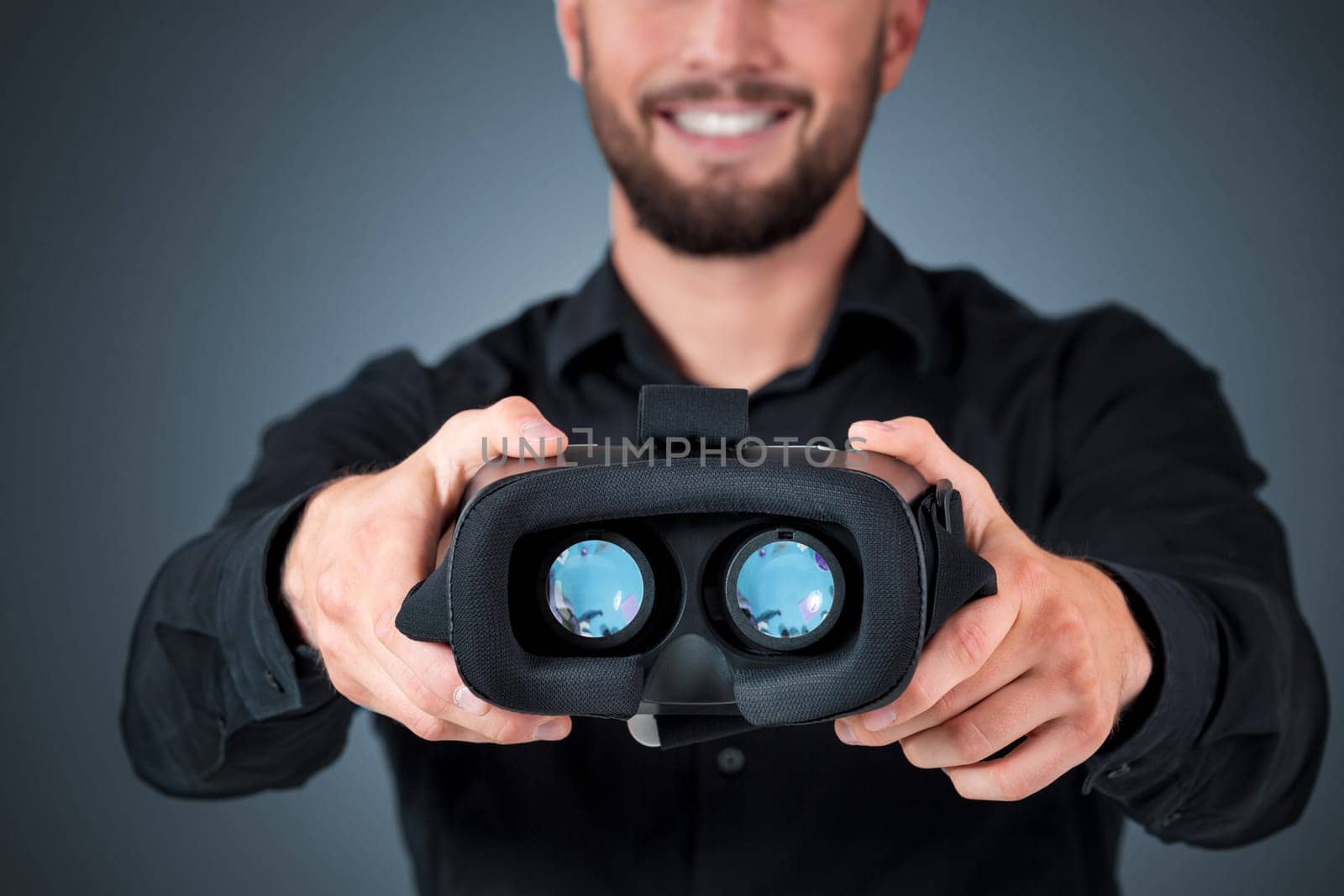 Excited young man using a VR headset and experiencing virtual reality on grey blue background. He holds glasses for viewer