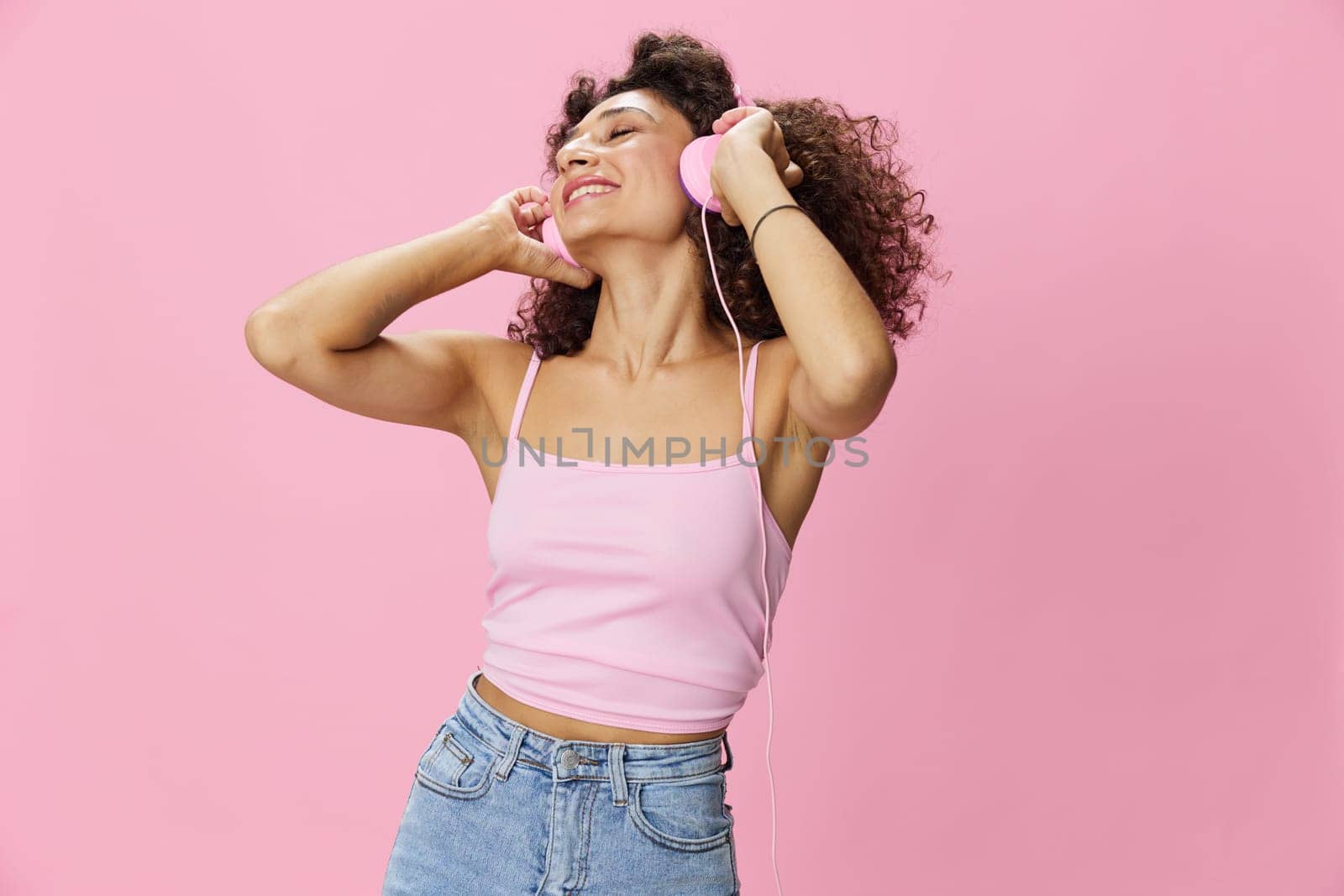 Happy woman wearing headphones with curly hair listening to music and dancing in a pink T-shirt and jeans on a pink background, copy space by SHOTPRIME