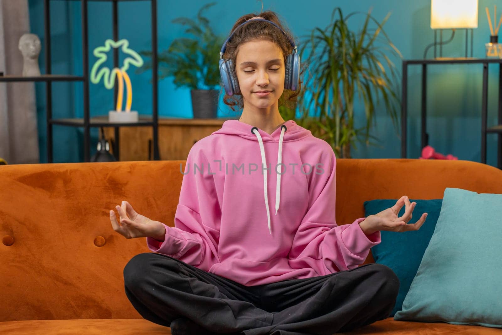 Keep calm down, relax. Preteen school girl breathes deeply with mudra gesture, eyes closed meditating with concentrated thoughts, peaceful mind. Young child kid at home room apartment sitting on couch