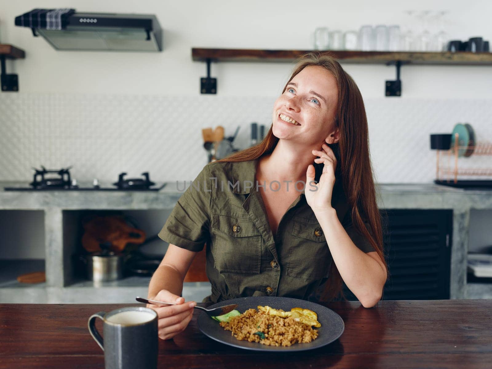 Woman with a smile eating Asian rice food in the kitchen at the wooden table, stylish interior in the background, lifestyle with phone in hand, blogger. High quality photo